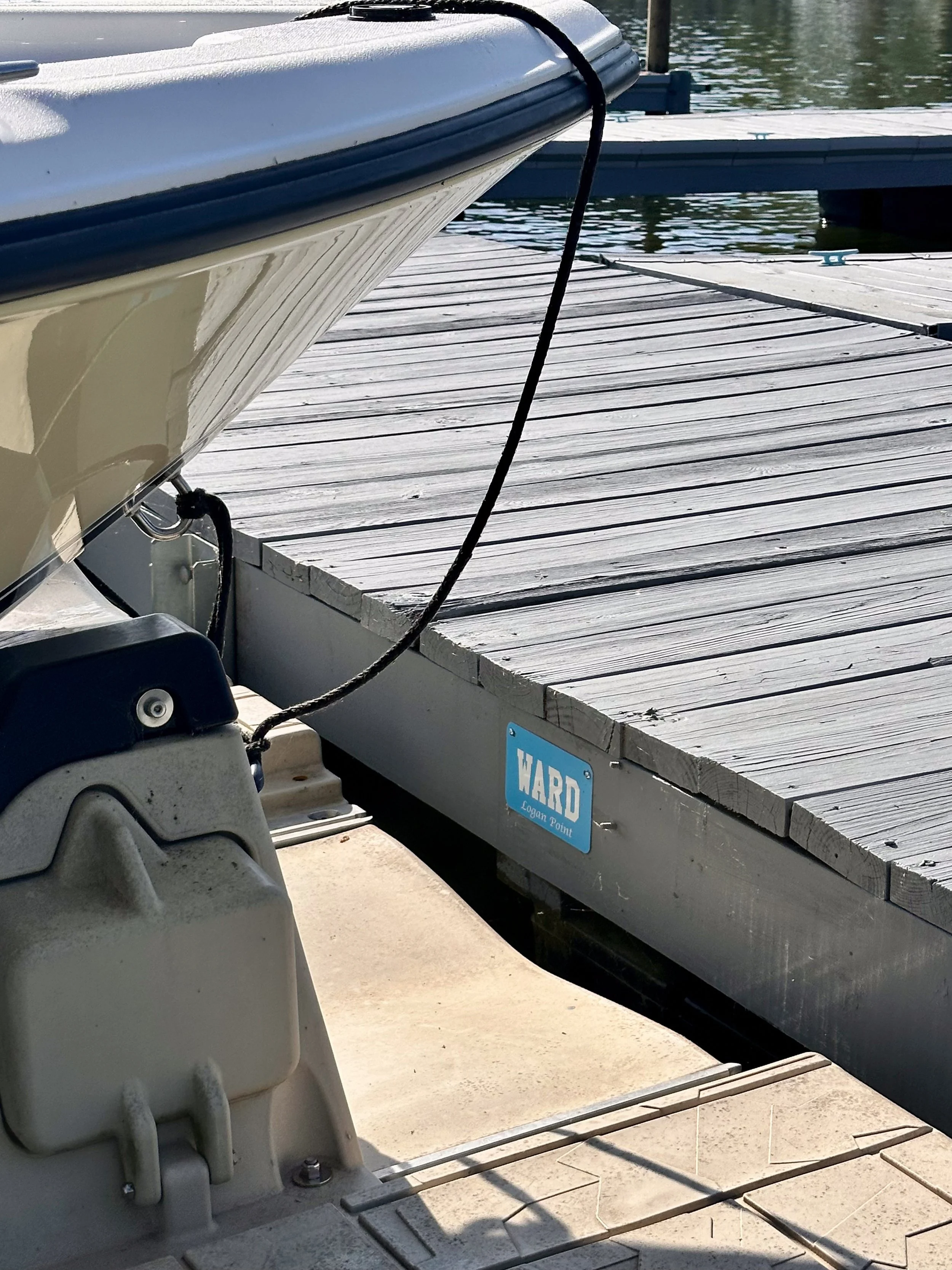 Close-up of a boat docked at a marina, showing part of the boat's hull, a dock cleat, a black rope, and a wooden dock with a blue sign that reads 'WARD'.