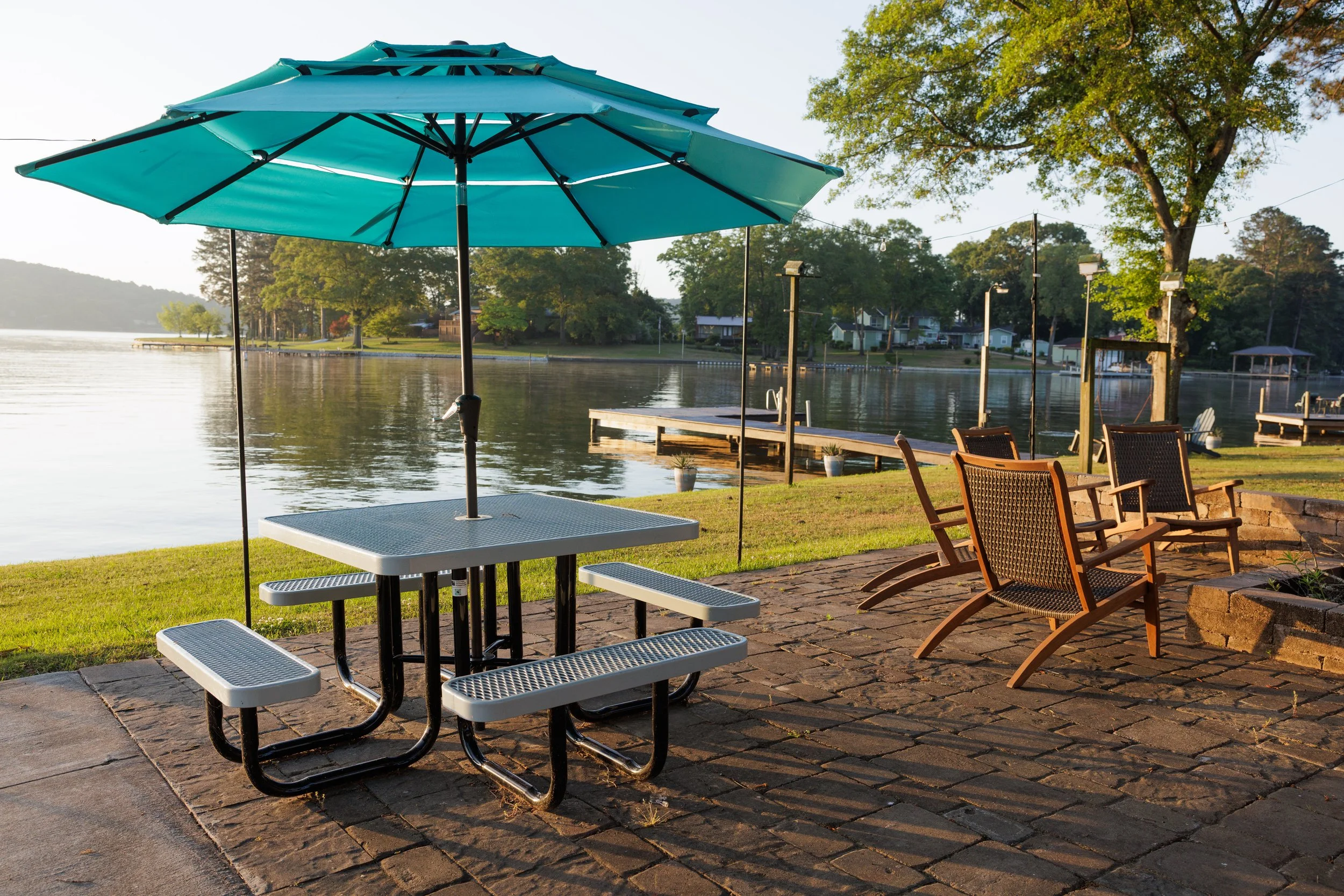 Outdoor patio by a calm lake with green trees, a blue umbrella over a picnic table, and lawn chairs on a brick-paved area during daytime.