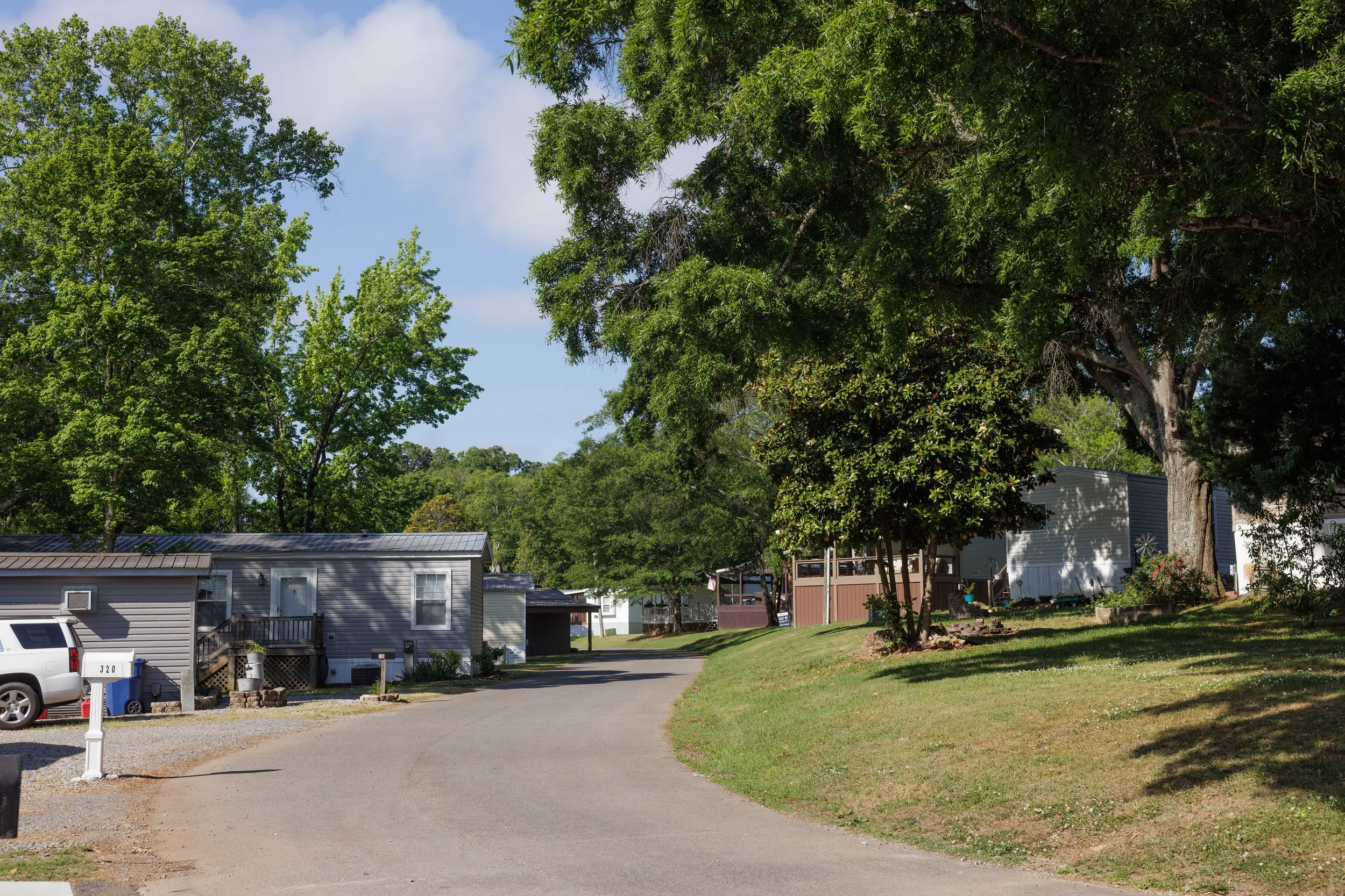 A residential neighborhood with mobile homes, trees, a driveway, and a parked car under a partly cloudy sky.