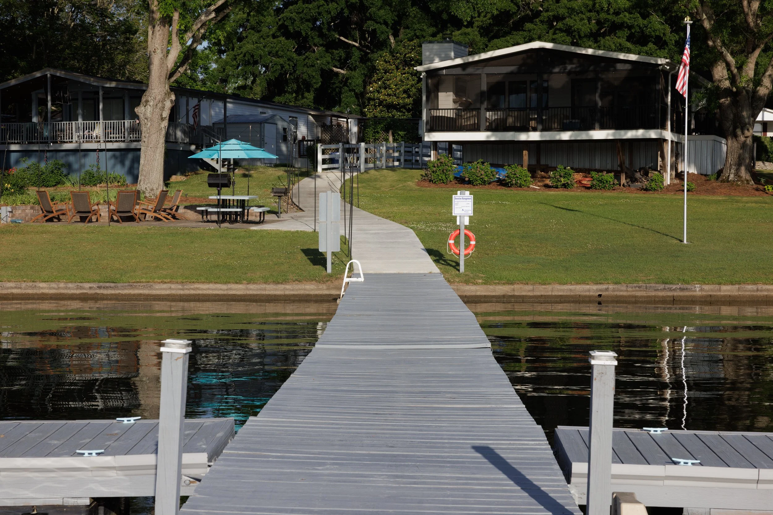 A wooden dock extending over a body of water towards a grassy yard with trees, a house, outdoor furniture, and an American flag.