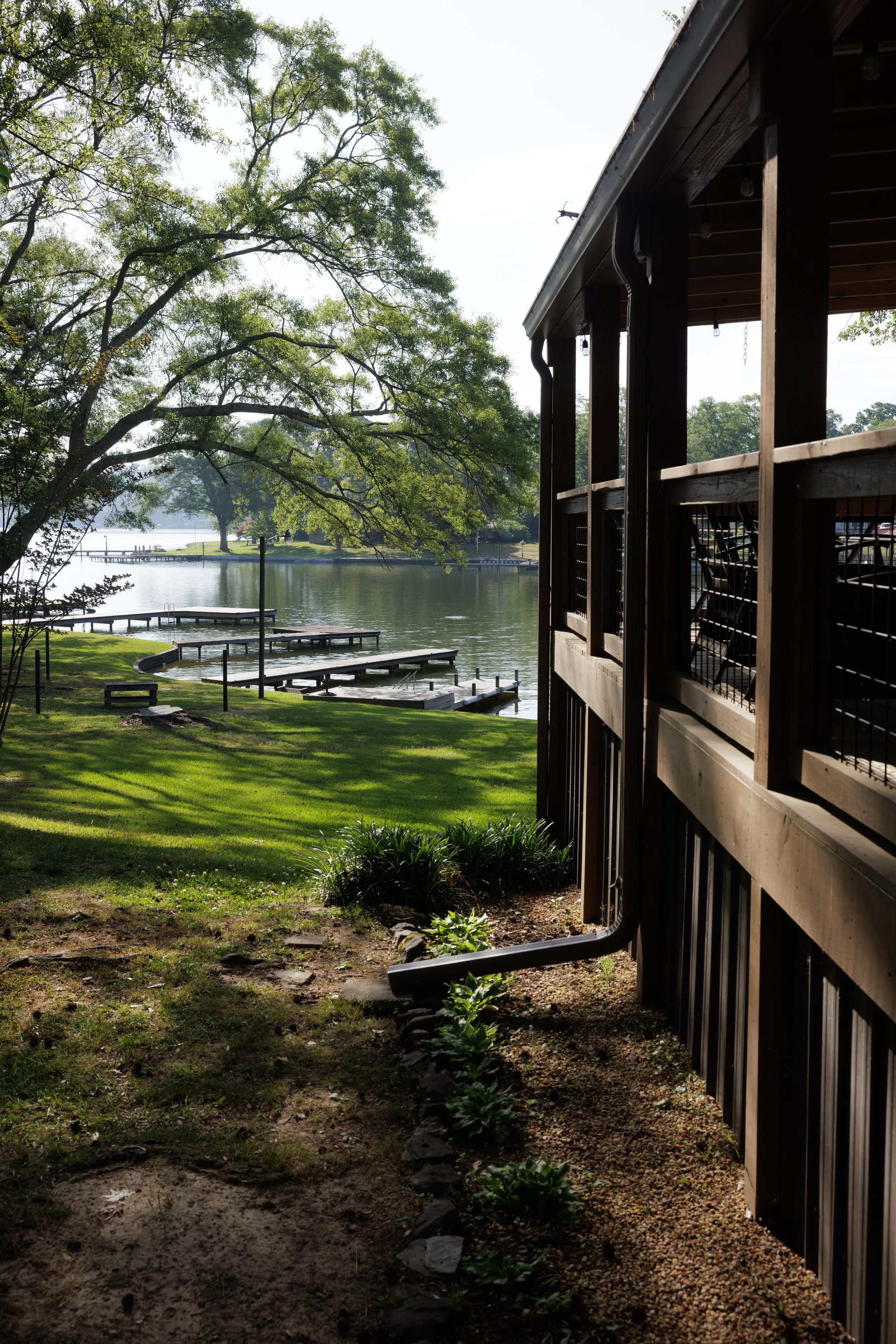 View of a lakeside area with grassy shore, trees, and a dock in the distance. Part of a wooden house or deck with support beams is visible on the right side of the image.