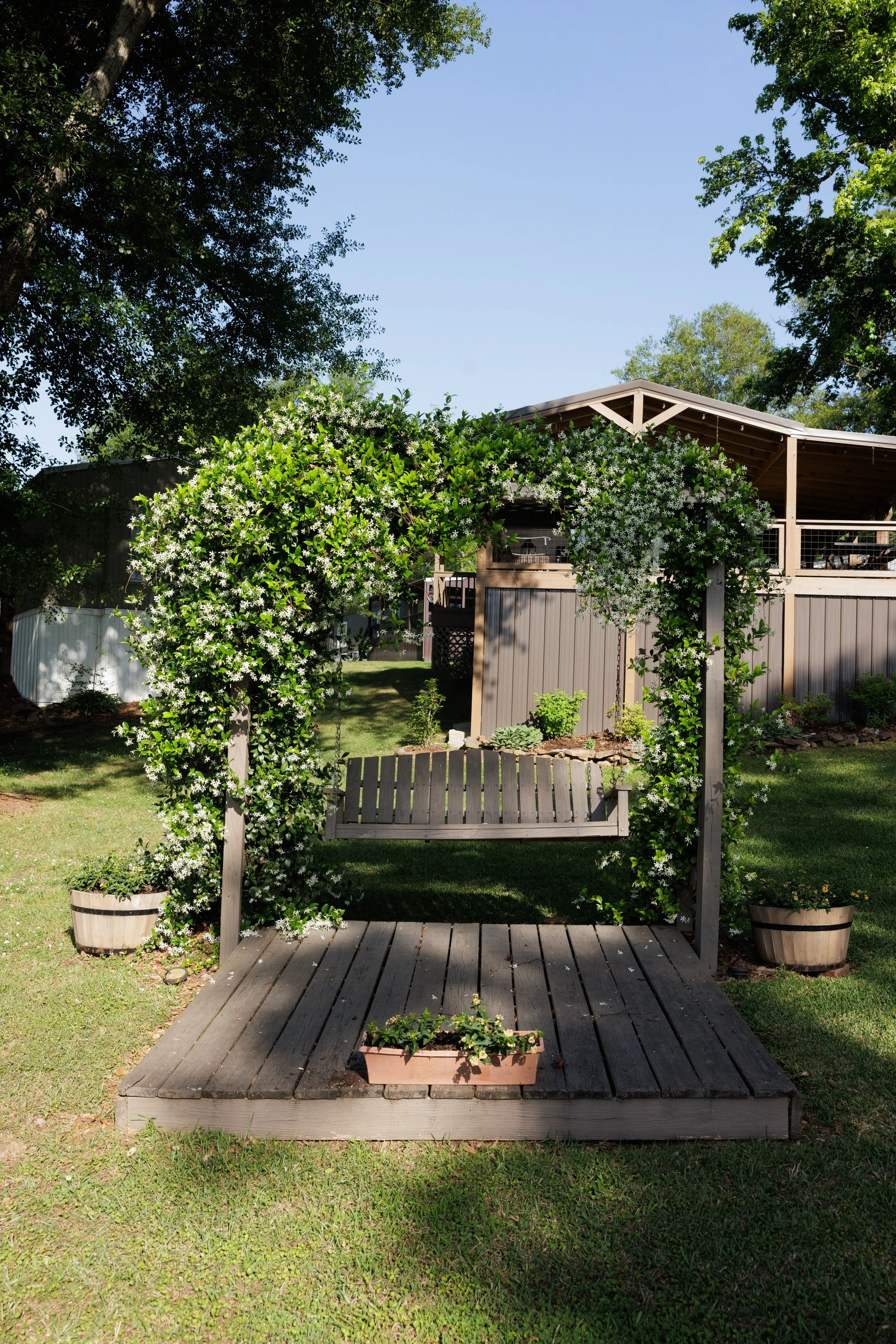 A backyard garden scene with a wooden patio, a garden swing in the center, and blooming vines on a pergola. Potted plants are placed on the ground, and a lush green lawn surrounds the area with tall trees in the background under a clear blue sky.