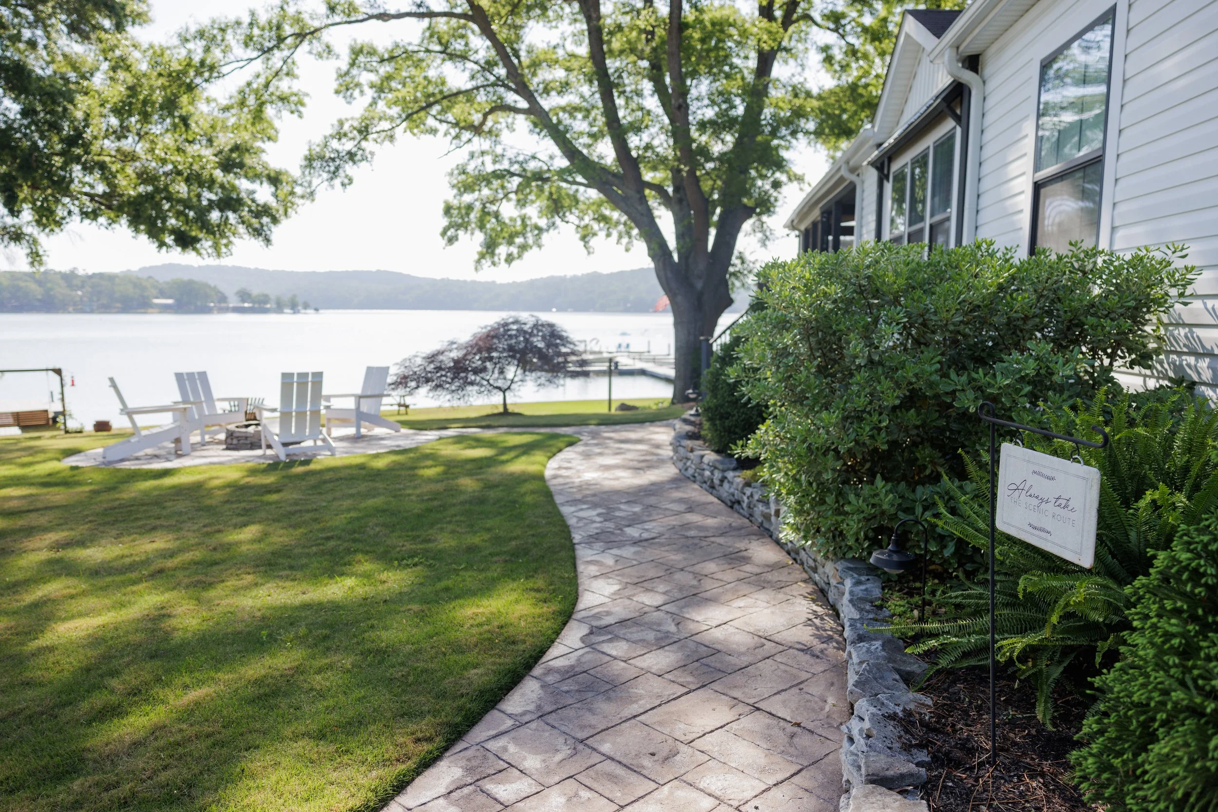 A lakeside scene with a paved walkway, green bushes, and trees, with white Adirondack chairs facing the water and a house with large windows on the right.