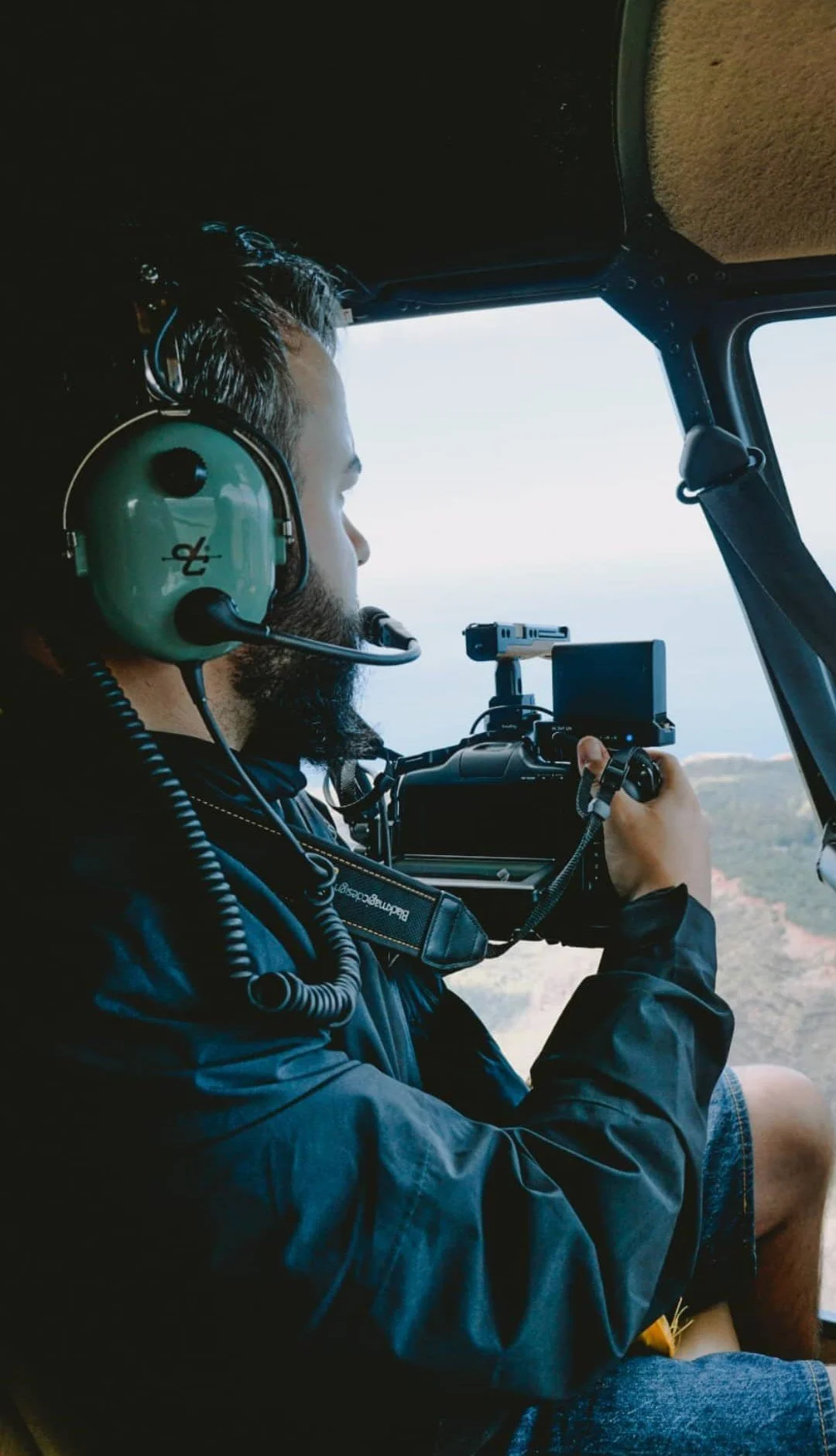Kevin Schie, Director of Photography, mit einem Kopfhörer und einem Filmkamera im Cockpit eines Hubschraubers, Blick nach vorne auf die Landschaft Hawaiis.
