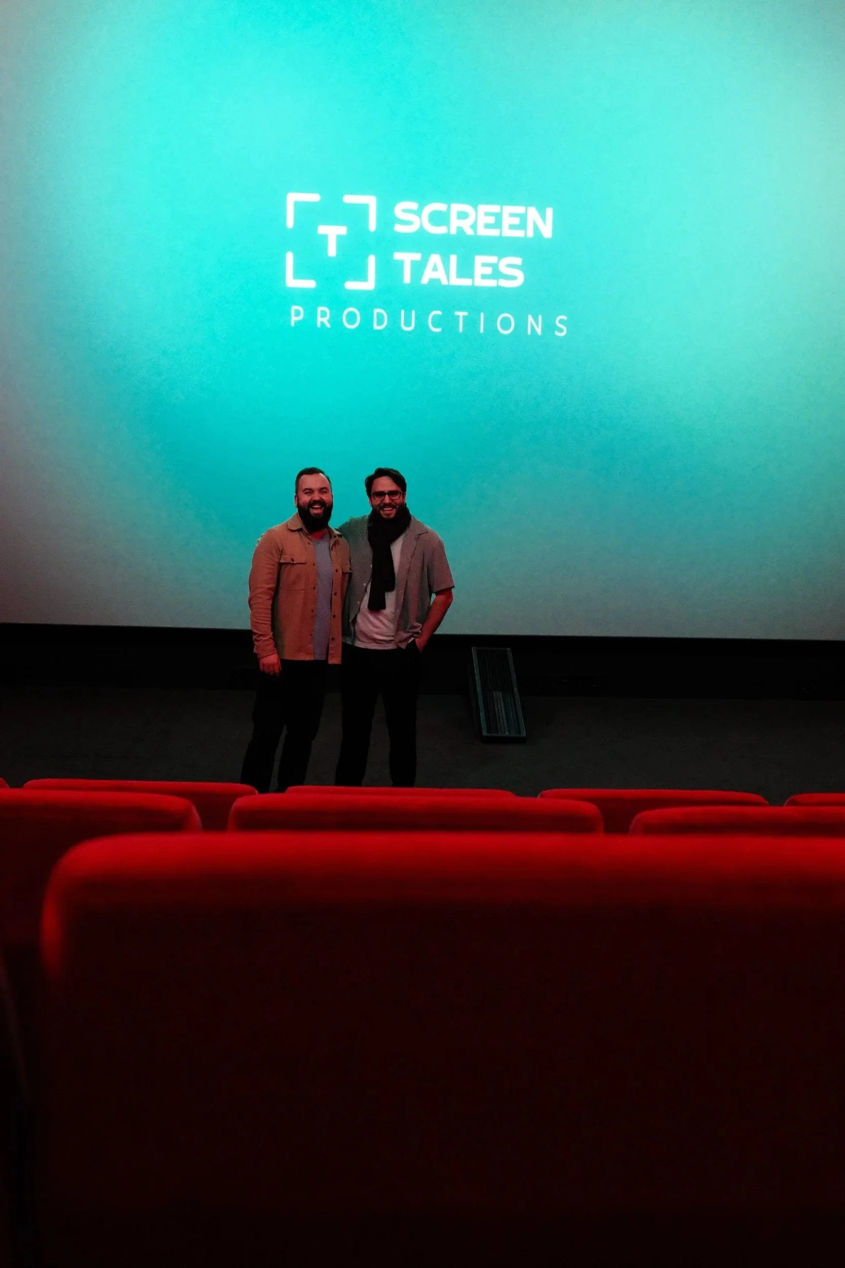Kevin Schie und RIccardo Lando stehen vor einer großen Leinwand mit dem Logo 'Tiny Screen Tales Productions' in einem Kino, leere rote Kinosaal-Stühle im Vordergrund.