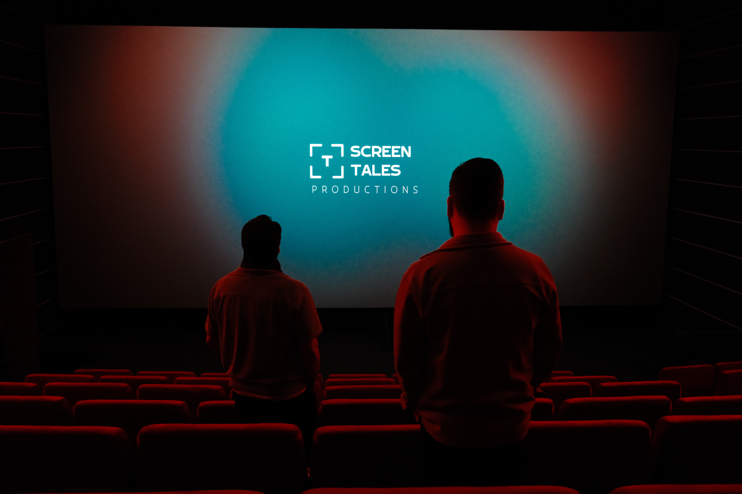 Riccardo Lando und Kevin Schie stehen in einem Kinosaal und schauen auf die große Leinwand, auf der das Logo von 'Tiny Screen Tales Productions' zu sehen ist.