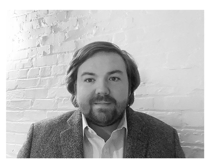 A man with medium-length hair and a beard, wearing a suit jacket and a collared shirt, sitting in front of a white brick wall, in a black and white photo.