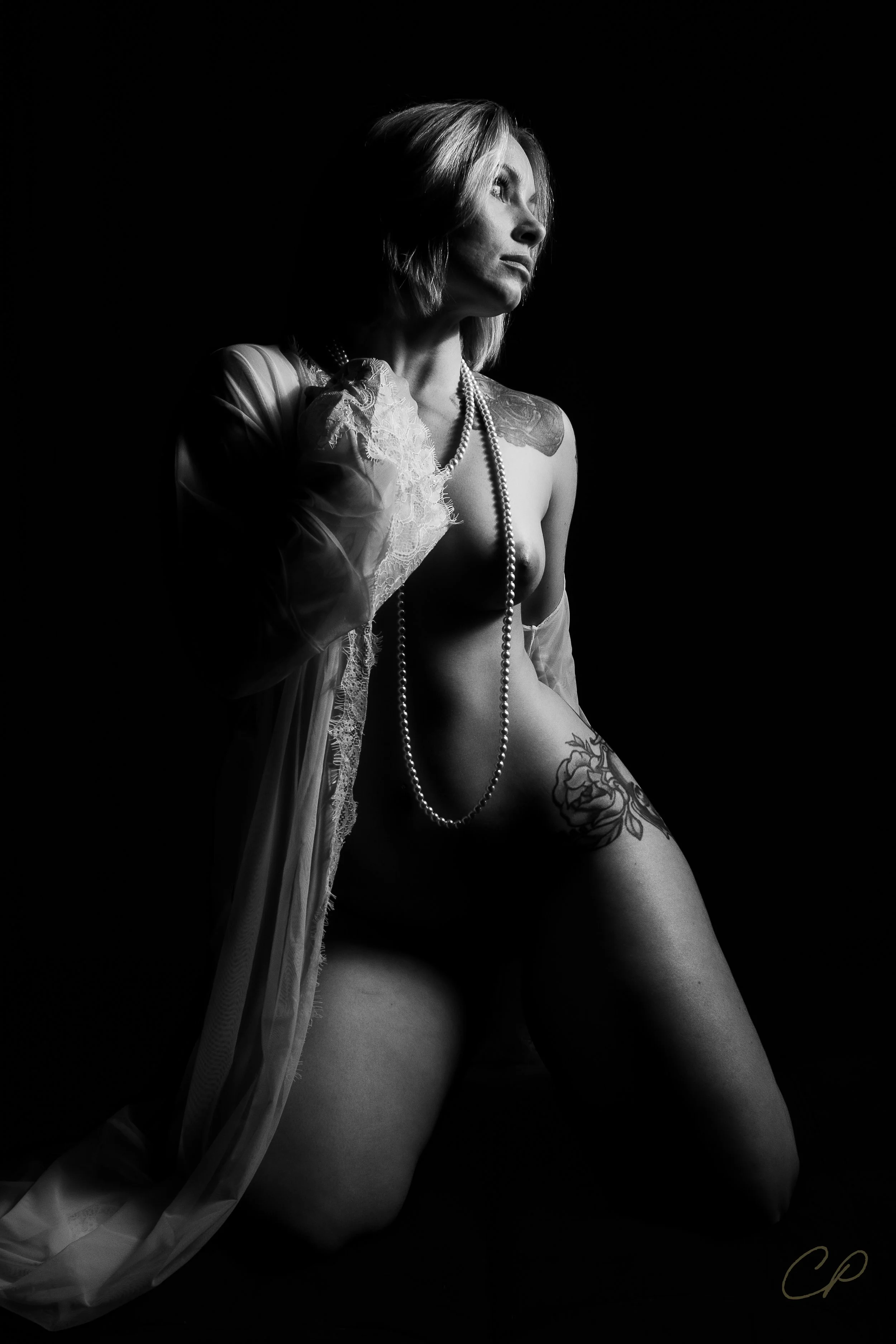 A black and white photograph of a kneeling woman with a flower tattoo on her thigh, wearing a pearl necklace and lace lingerie, with one hand touching her face, illuminated by side lighting against a dark background.