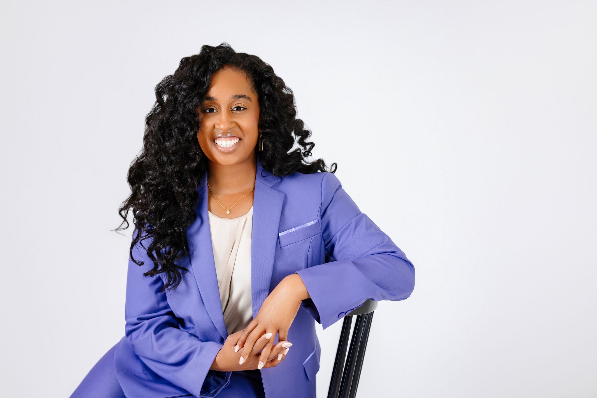 A woman with long black curly hair wearing a purple blazer and cream blouse, smiling and sitting on a chair against a plain white background.