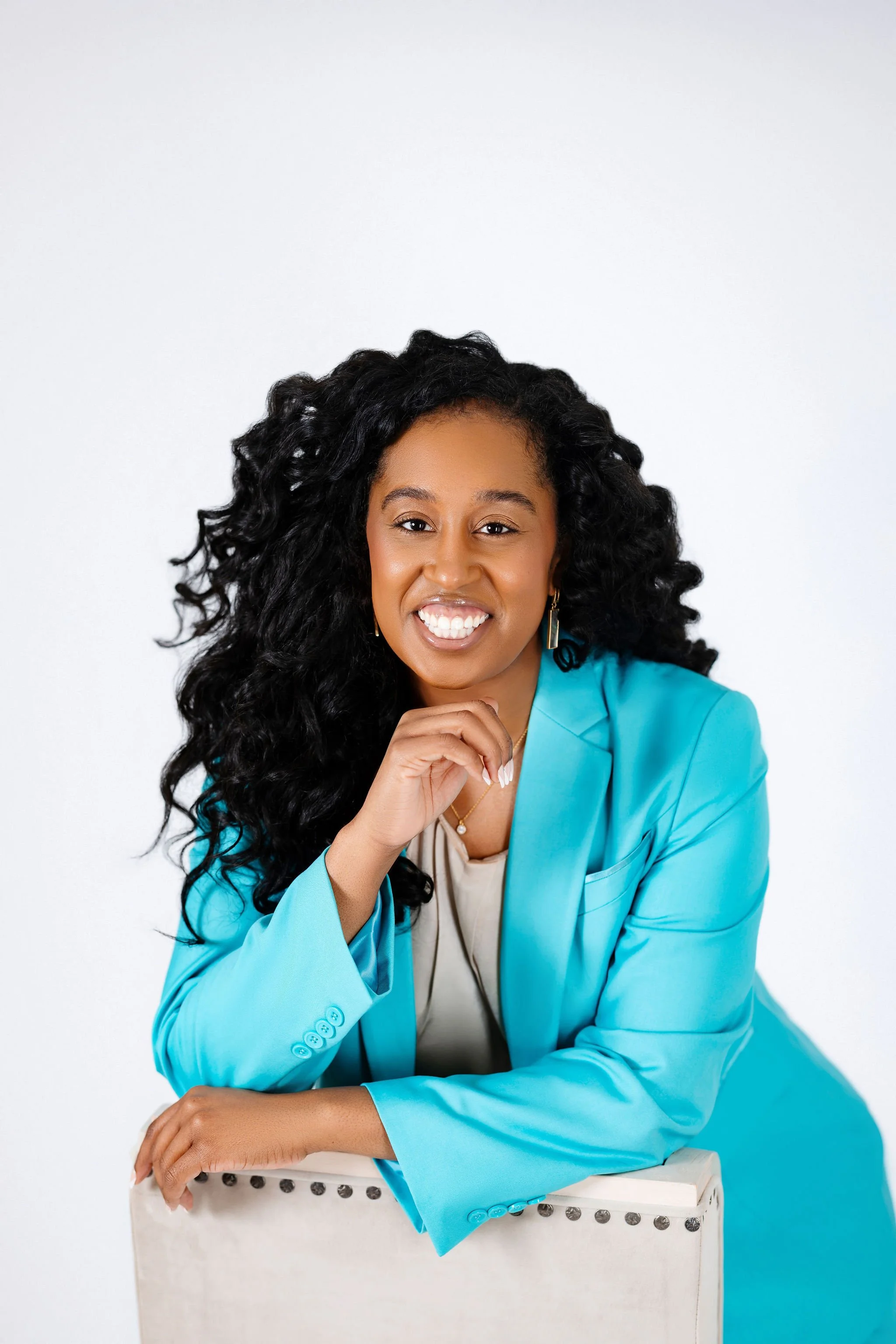 A woman with curly black hair wearing a bright blue blazer, beige top, and gold jewelry, smiling and resting her chin on her hand against a white background.