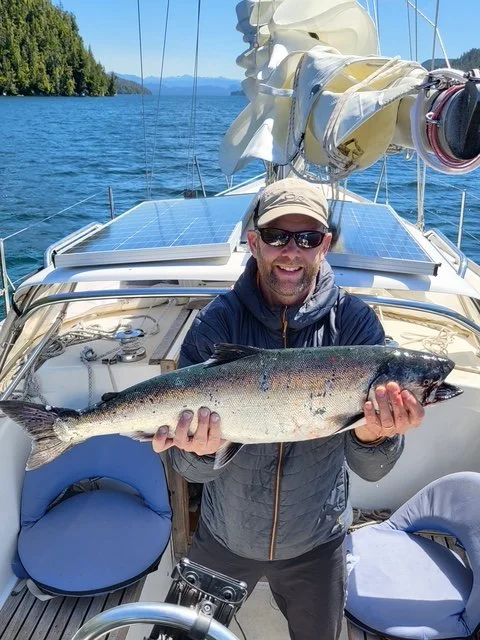Man wearing sunglasses, a cap, and a gray jacket holding a large salmon caught using glow-in-the-dark fishing tackle.