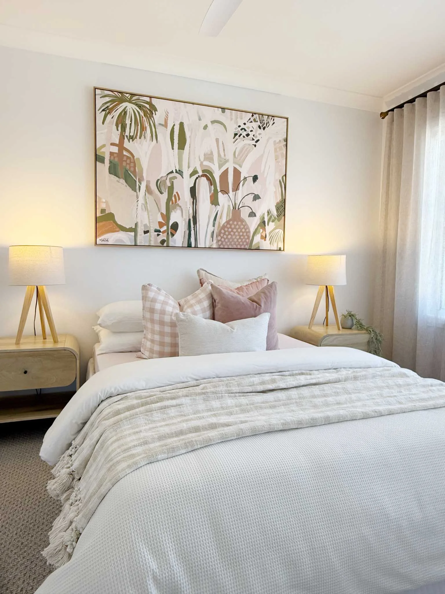 A bedroom with a neatly made bed, featuring a white textured blanket and multiple pillows in soft pastel shades and patterns. There are two matching wooden nightstands with lamps, a small plant on one, and a large framed abstract botanical painting above the bed. Cream-colored curtains cover the window to the right.