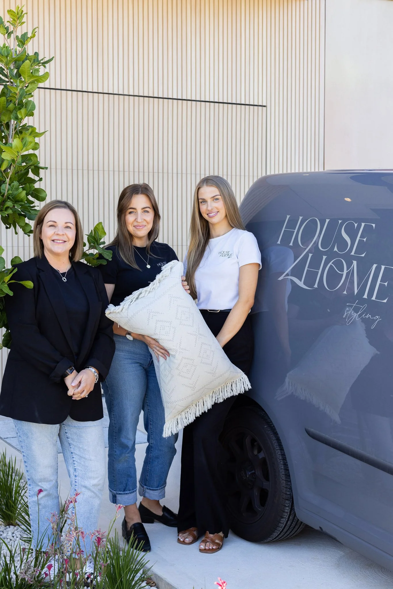 Three women standing outdoors near a gray house and a gray vehicle with the words "House 2 Home" on it, smiling and holding a decorative pillow.