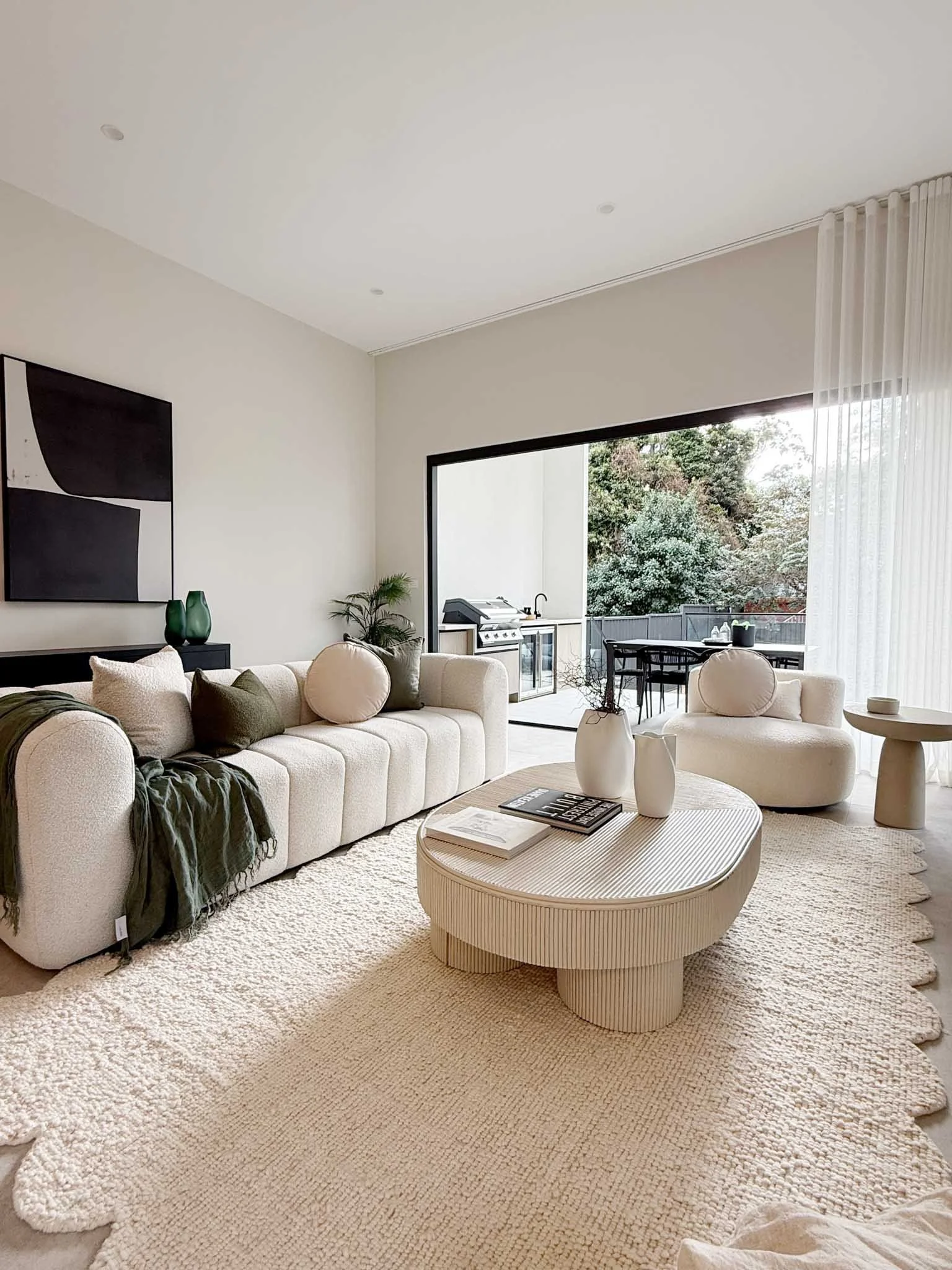 Modern living room with a beige sofa and chairs, a coffee table with decorative vases, a textured cream rug, and an open balcony with outdoor seating and greenery.