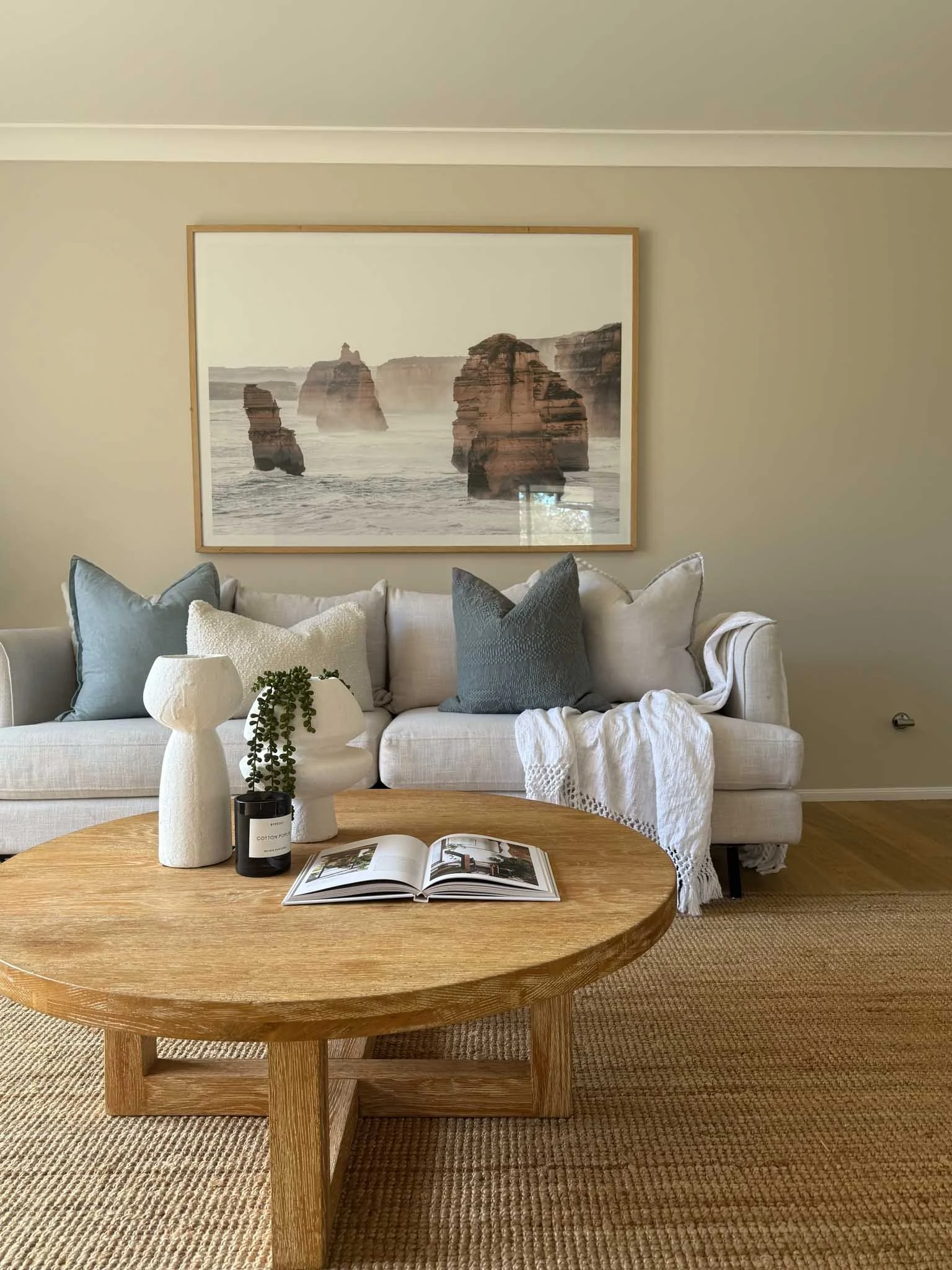 Living room with a white sofa, blue and white pillows, a wooden coffee table with vases and an open book, a framed ocean landscape painting on the wall, neutral wall color, and a beige rug.