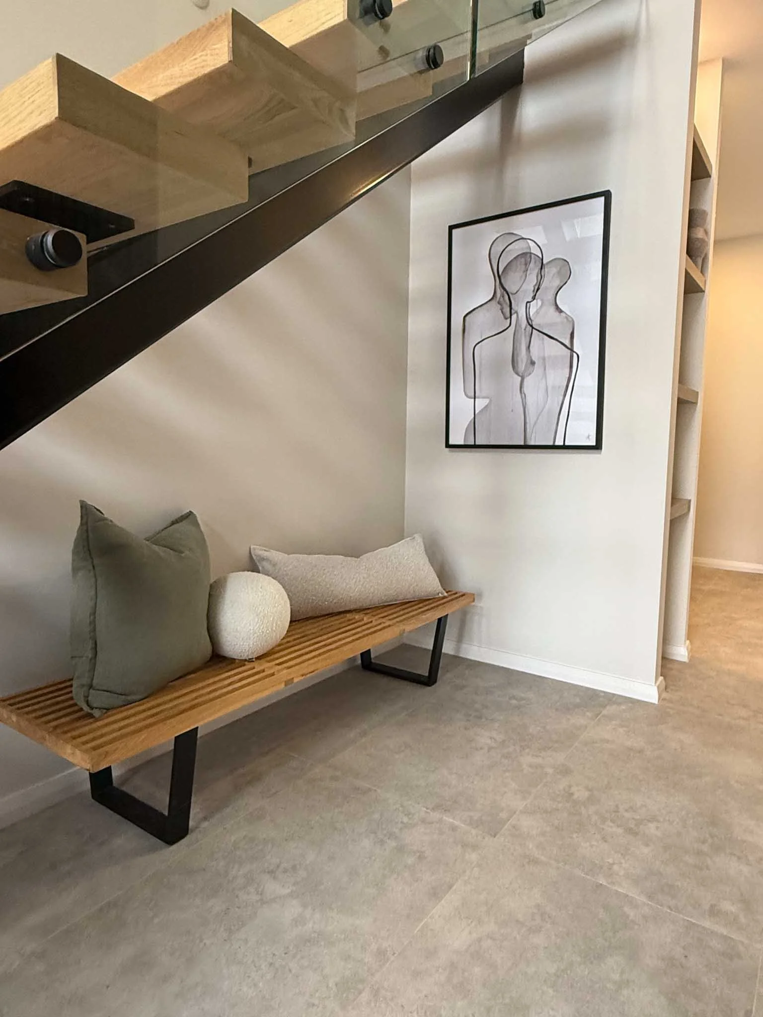 A wooden bench with three pillows and a decorative round object, placed underneath a modern staircase with wooden steps and glass railing, in a minimalist interior with a black and white line art portrait on the wall.