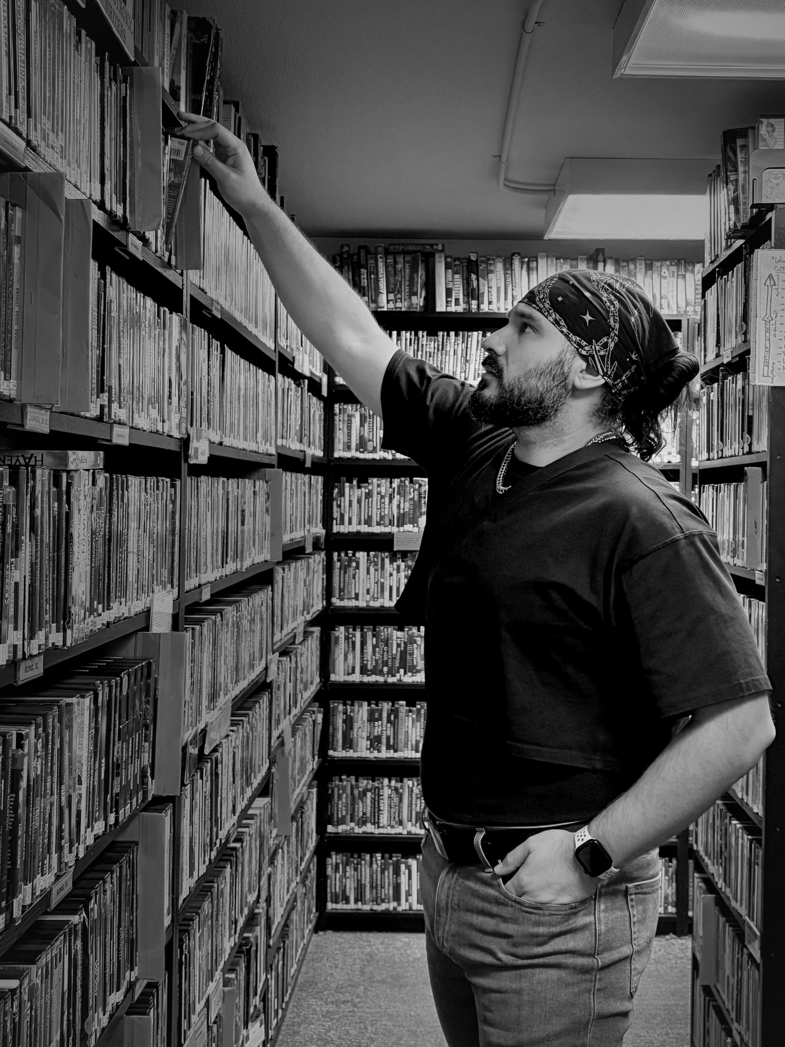 A man in a black t-shirt and jeans is reaching for a book on high shelf in a library, wearing a patterned bandana and a smartwatch.