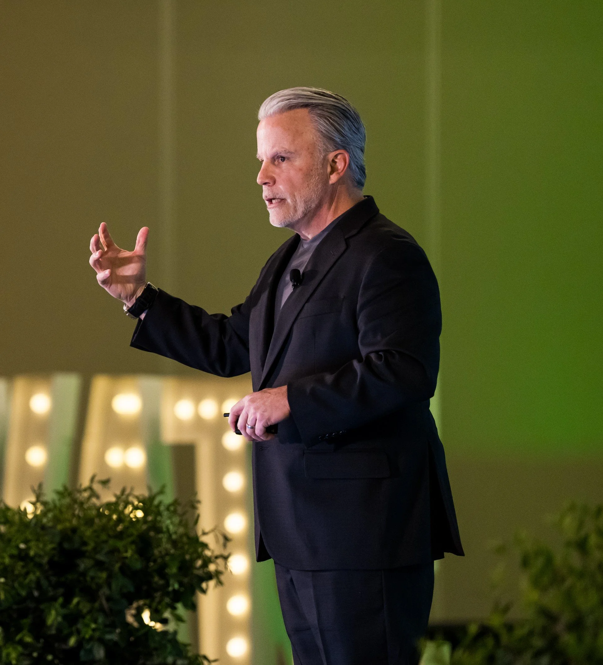 A man with gray hair and beard, wearing a black blazer and gray shirt, giving a presentation with a green background and illuminated letters.