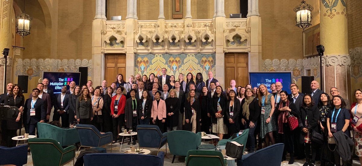 A large group of people posing for a photo in a decorated, historic-style room with ornate architecture. There are two large screens displaying 'The Multilevel Effect' on either side of the group, and several chairs and tables in the foreground.