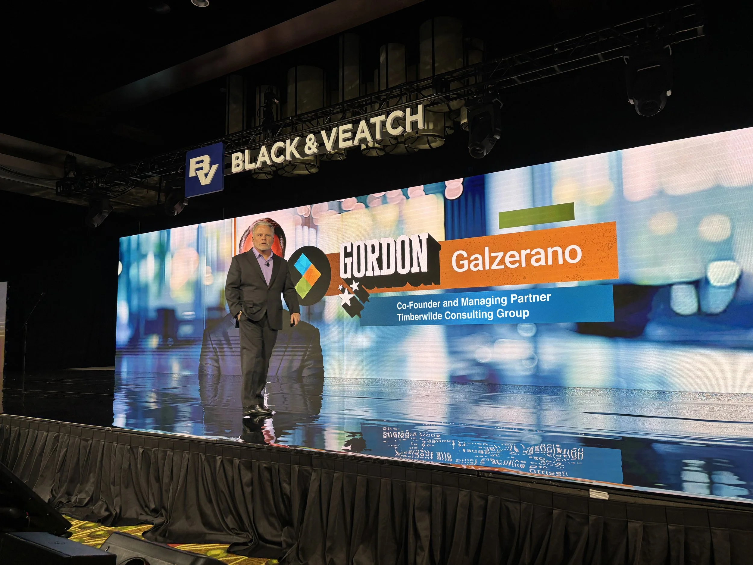A man in a dark suit and light shirt stands on a stage with a large digital screen behind him, displaying his name Gordon Galzerano, his title, and the company Timberwilde Consulting Group. The stage is part of an event at Black & Veatch, with a sign overhead displaying the company name.