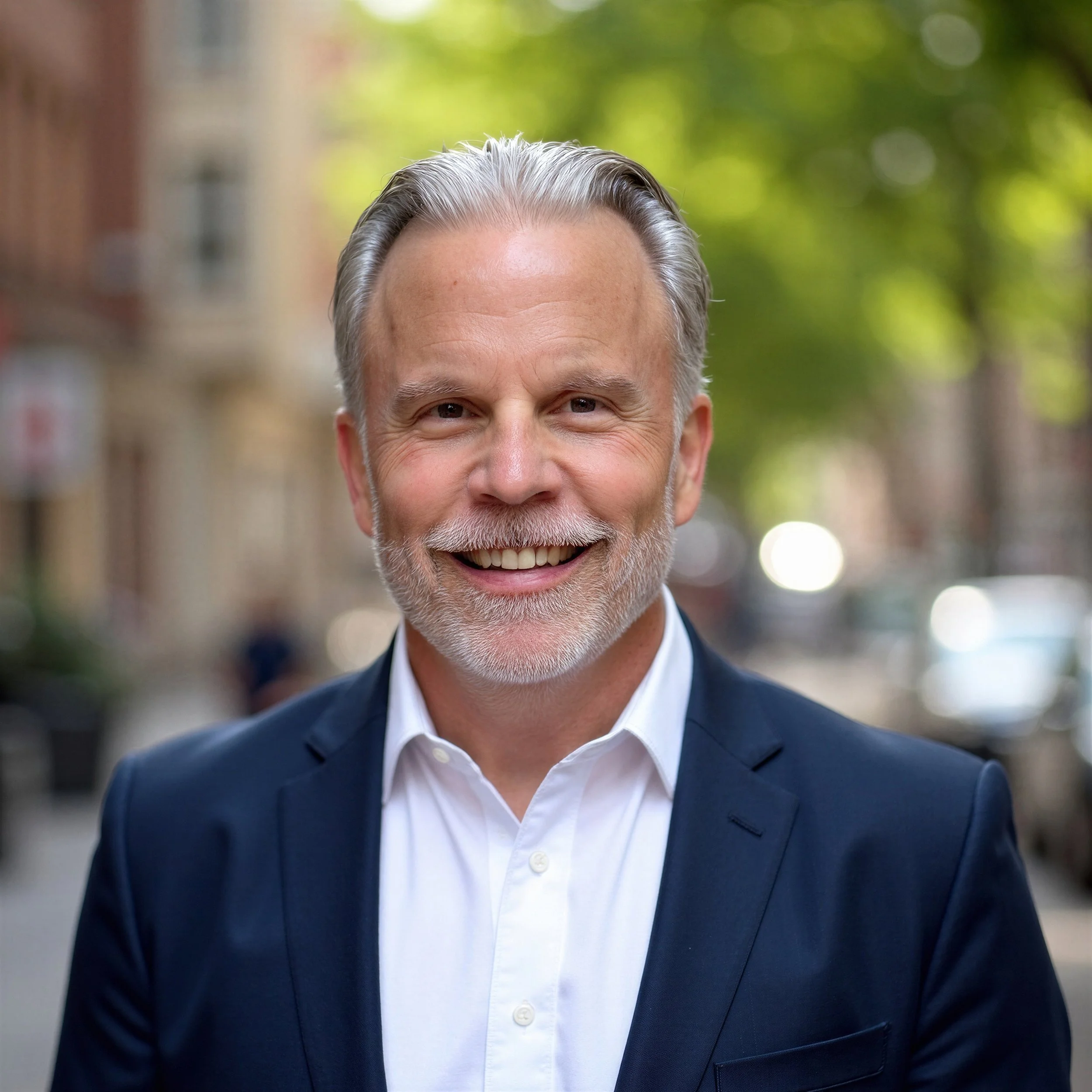 Close-up of Gordon Galzerano, wearing a dark suit and white shirt, standing outdoors on a city street with blurred background of buildings and trees.