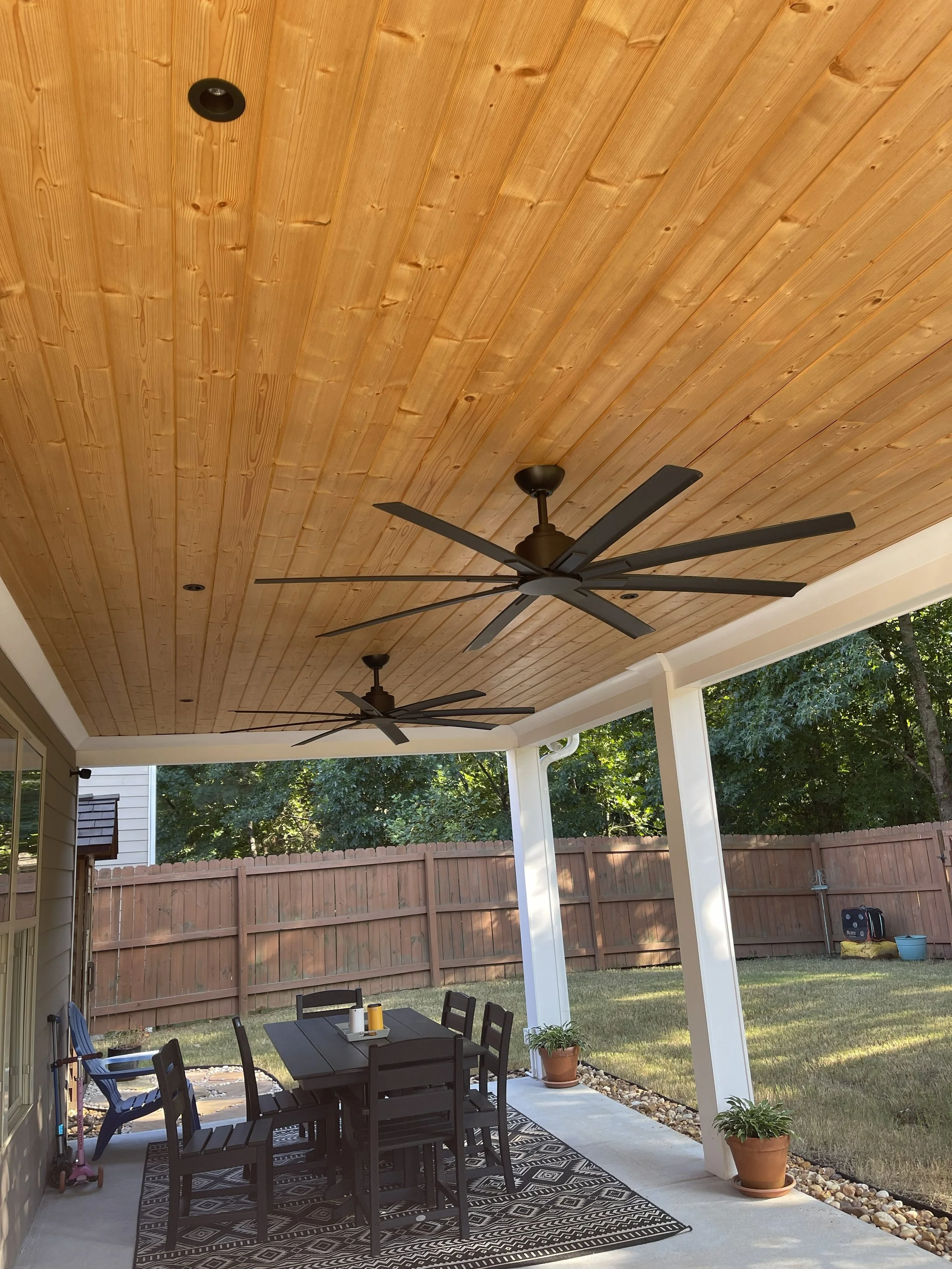 Covered outdoor patio with a wooden ceiling, two ceiling fans, and a patio table with six chairs, along with potted plants and a garden in the background.