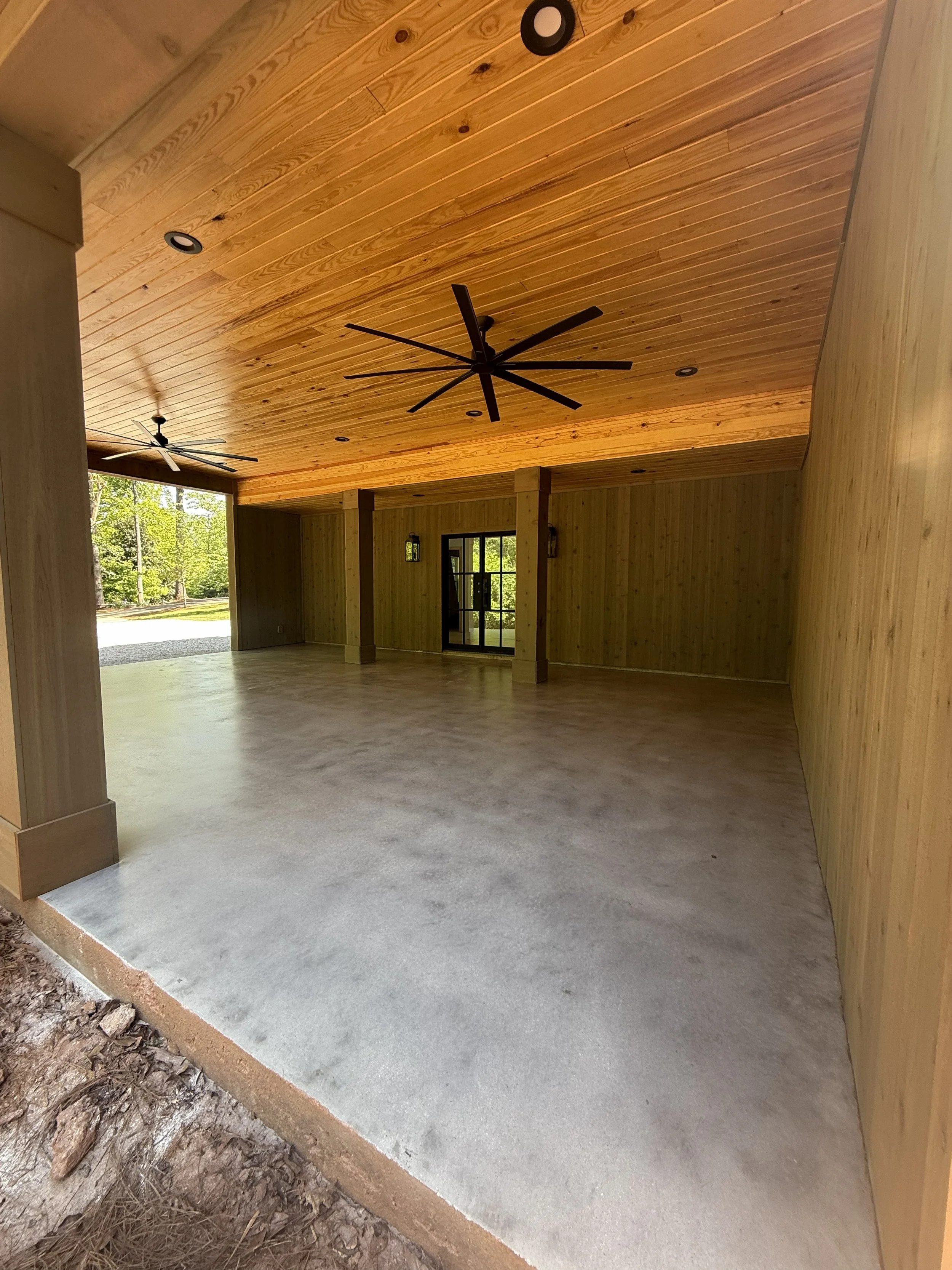 Empty carport with wood-paneled ceiling, concrete floor, and two ceiling fans, overlooking a wooded outdoor area.