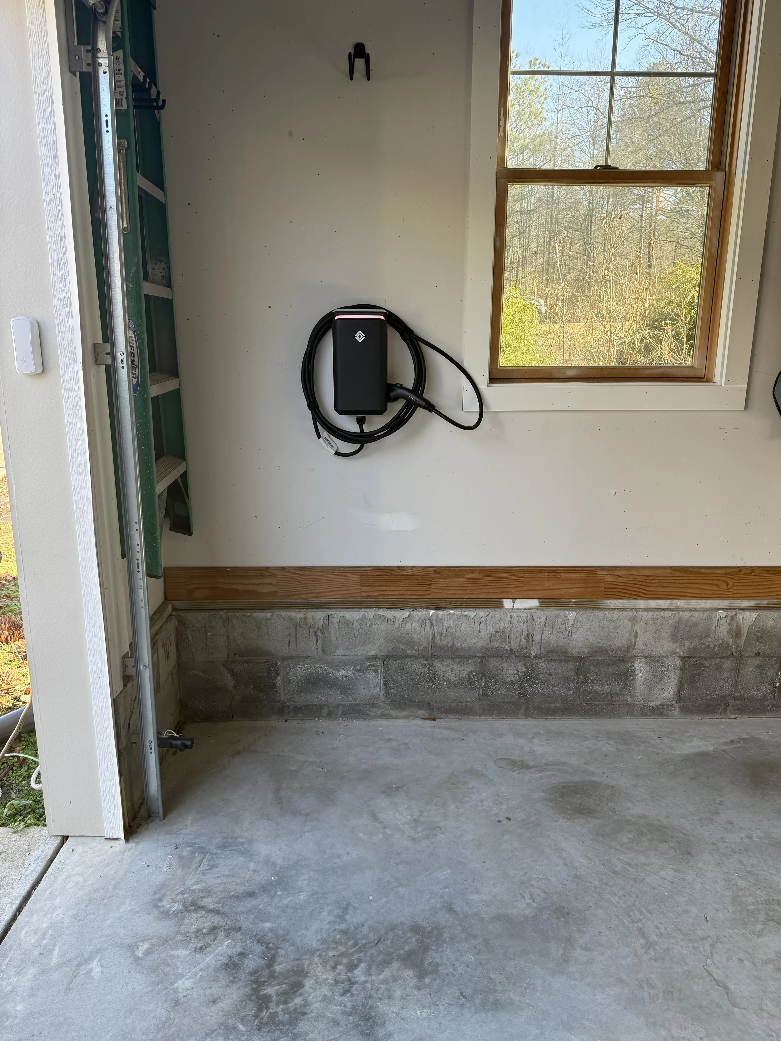 Empty garage with concrete floor, a window showing trees outside, a wall-mounted electric vehicle charging station, and a ladder on the left.