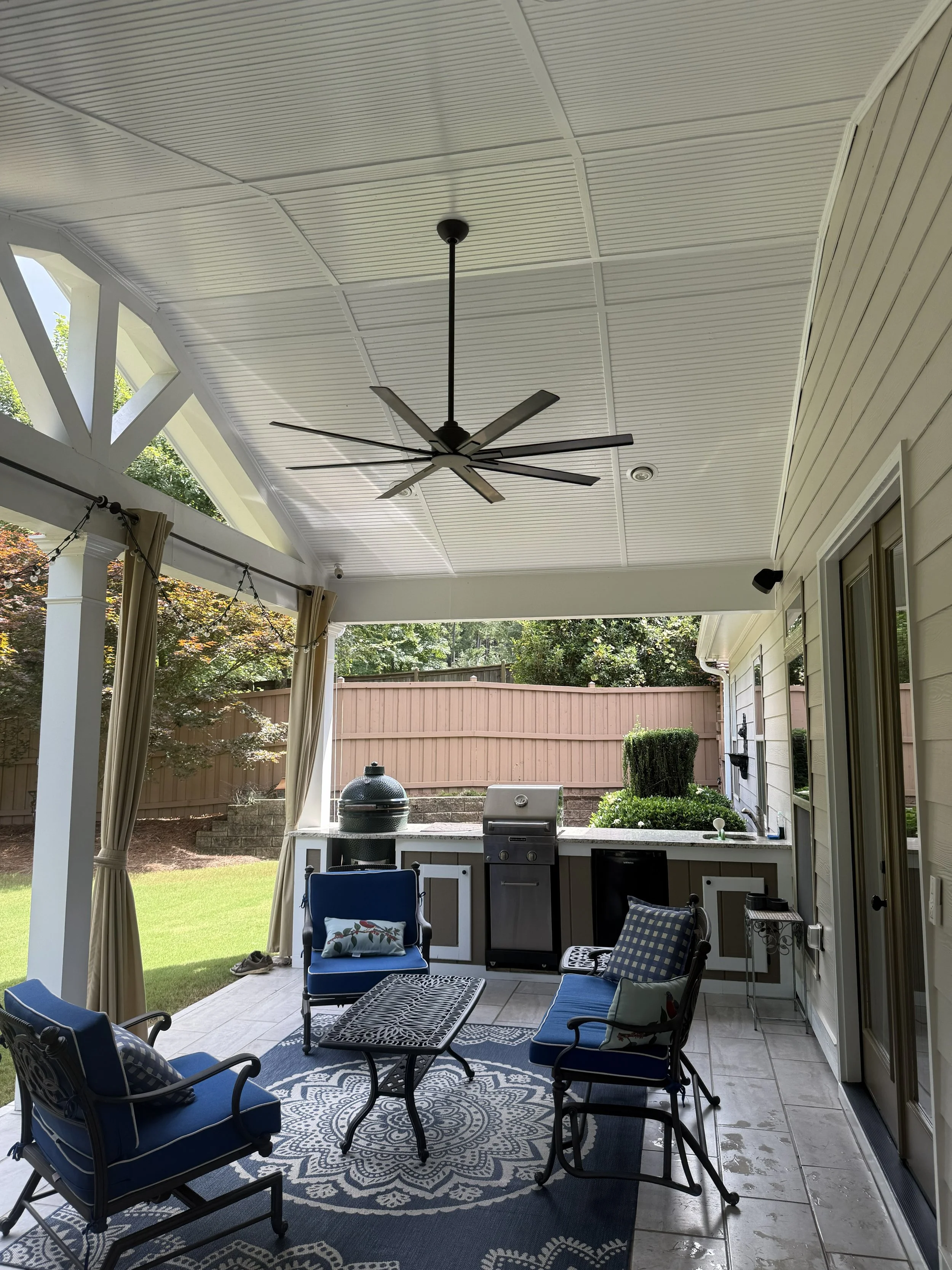 Covered outdoor patio with ceiling fan, ceiling with white panels, blue cushioned chairs, small table, outdoor grill, and a decorative rug, surrounded by a lawn and fencing.