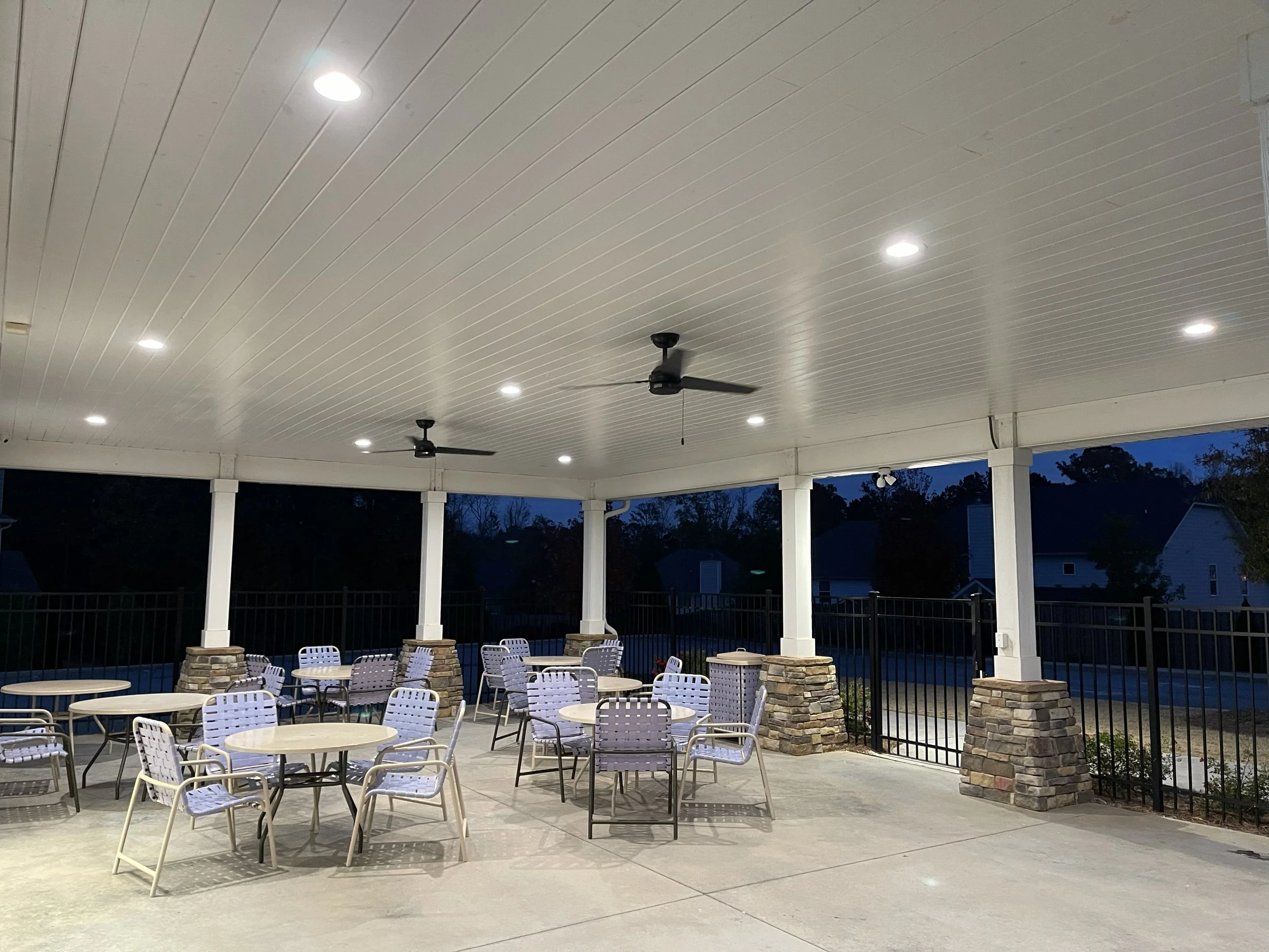An outdoor covered patio with round tables and chairs, ceiling fans, and recessed lighting, with a black metal fence and trees in the background.