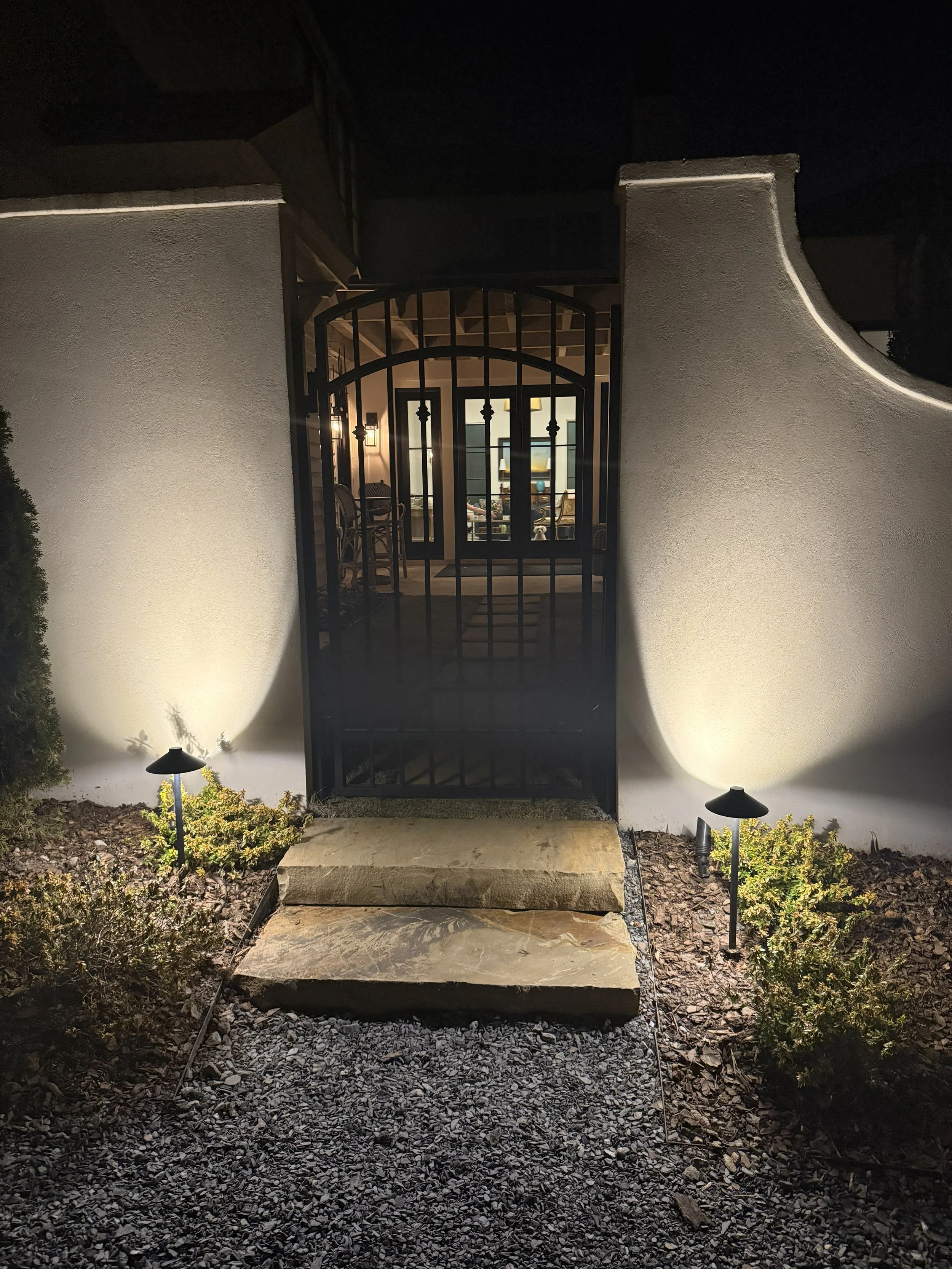 Nighttime view of a house entrance with a black metal gate, stone steps, and exterior lighting illuminating small bushes and gravel ground.