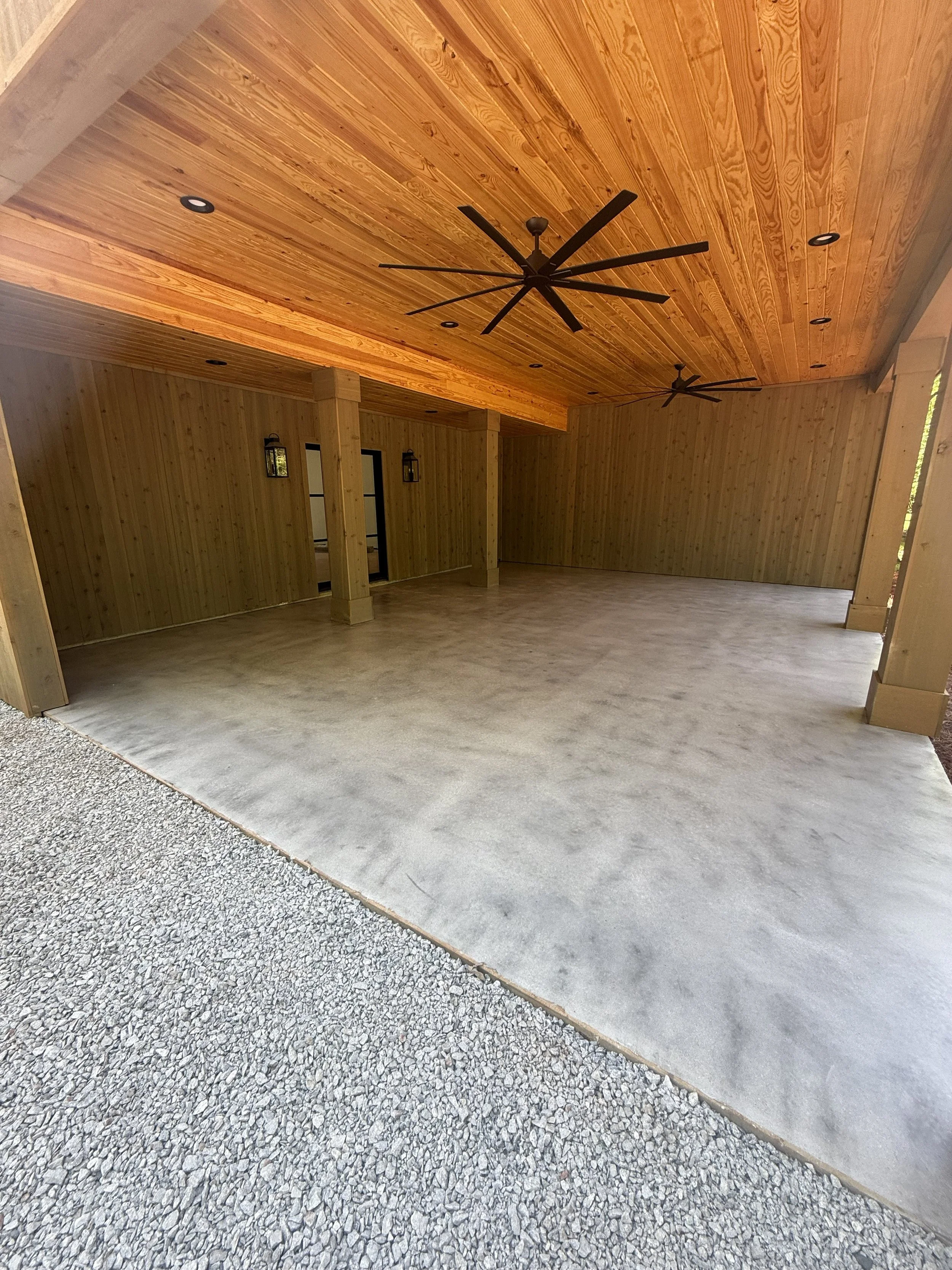 Empty wooden garage with a concrete floor, wooden ceiling, and ceiling fans, illuminated by natural light.