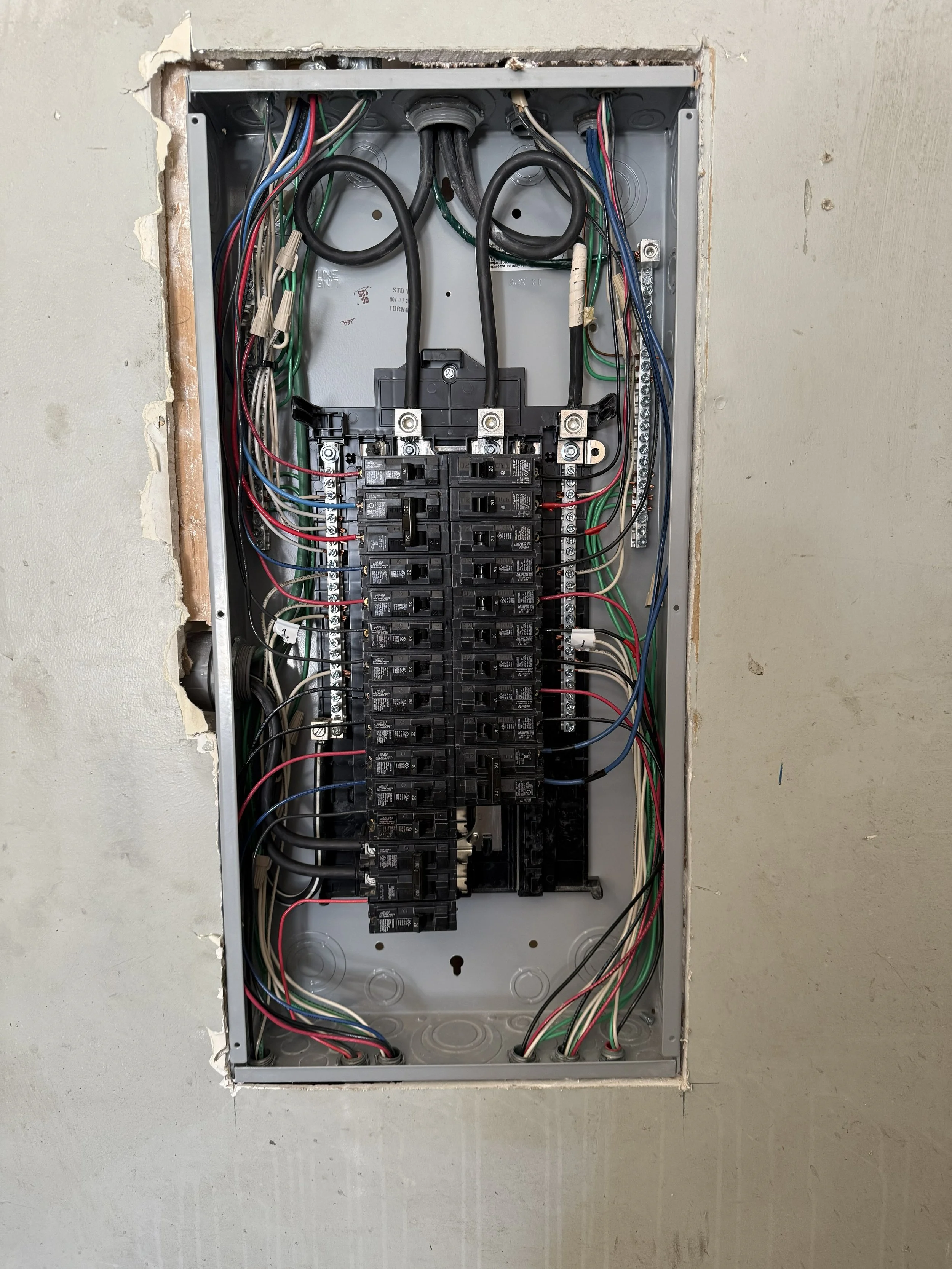 Open electrical breaker box with multiple circuit breakers and wiring inside a wall.