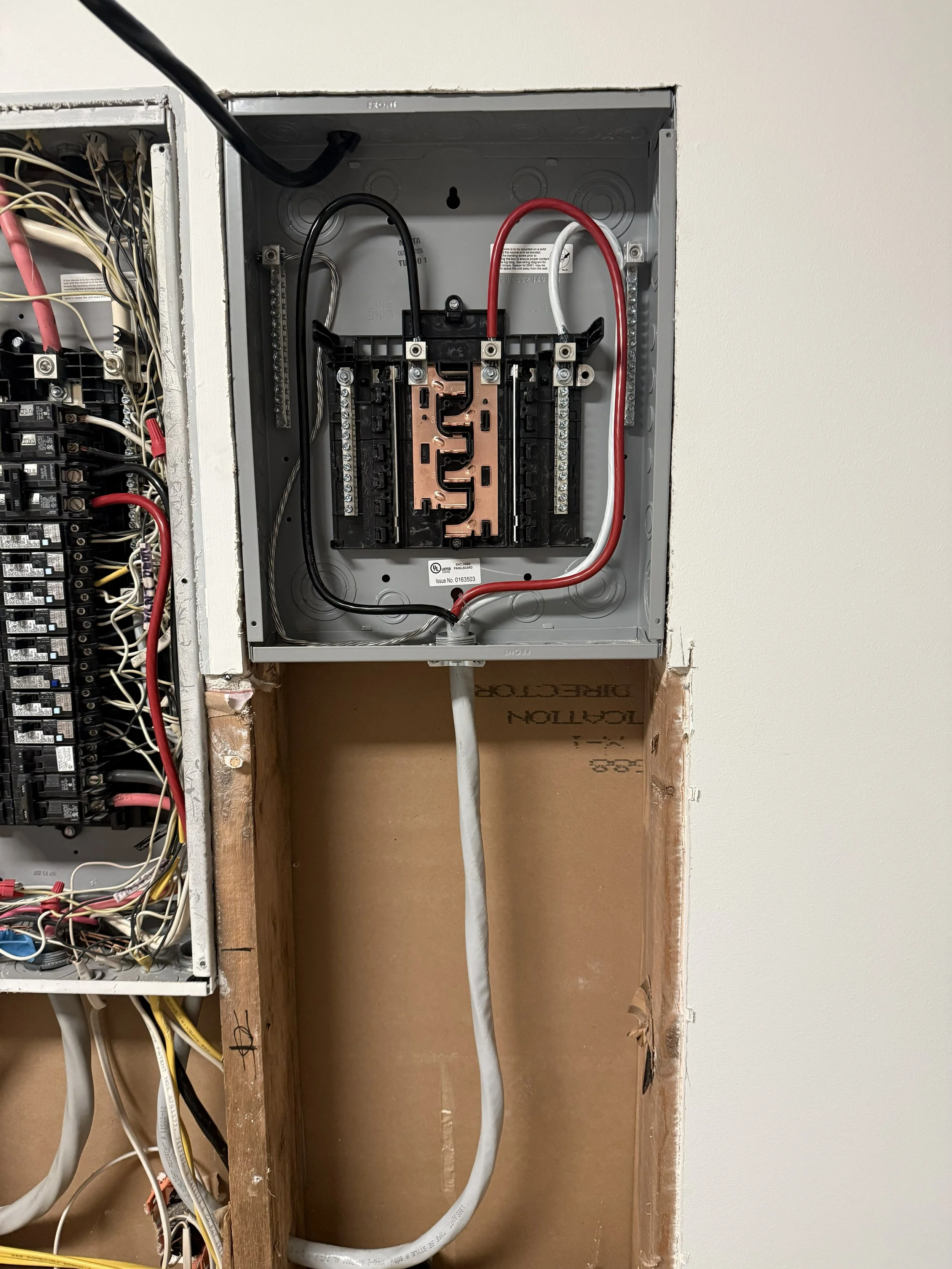 An electrical panel with wiring, circuit breakers, and a copper coil inside a metal box, with some parts not fully installed, on a wall with exposed drywall.