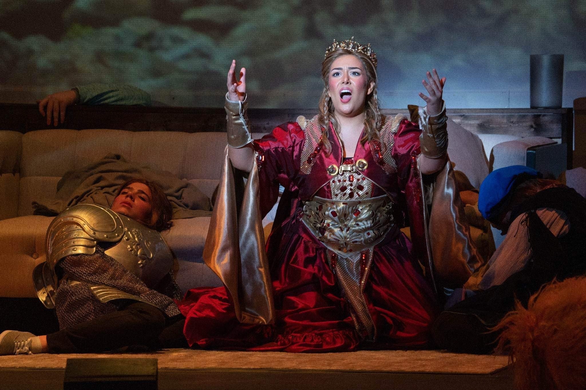 A woman dressed as a queen, wearing a red and gold costume with a crown, kneeling on the floor with her arms raised and mouth open, appearing to be in distress or making a dramatic plea. There are two children dressed as knights lying on the floor be