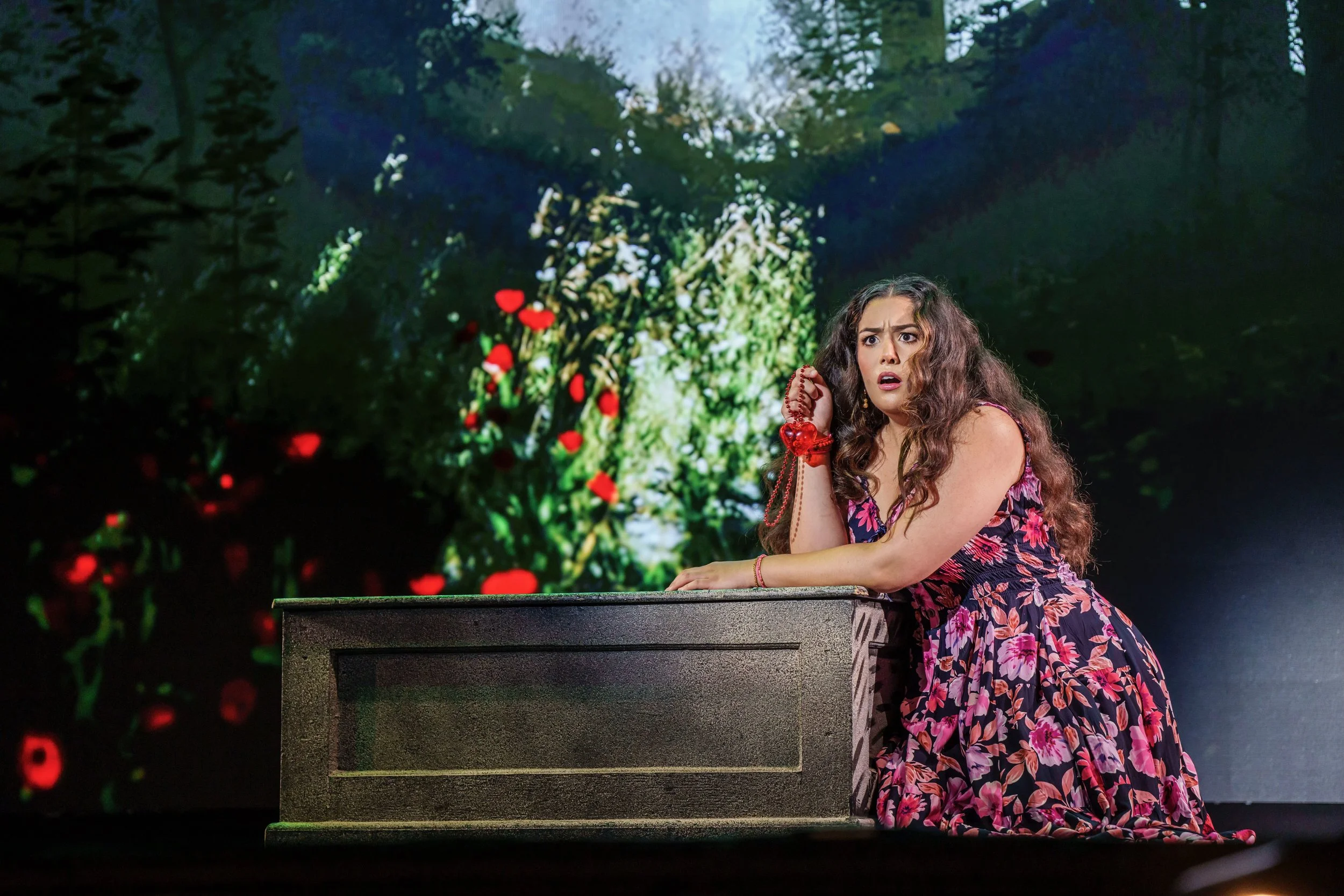 A woman with long curly hair in a floral dress, kneeling and leaning on a black box with a surprised or worried expression, set against a colorful background of trees and flowers.