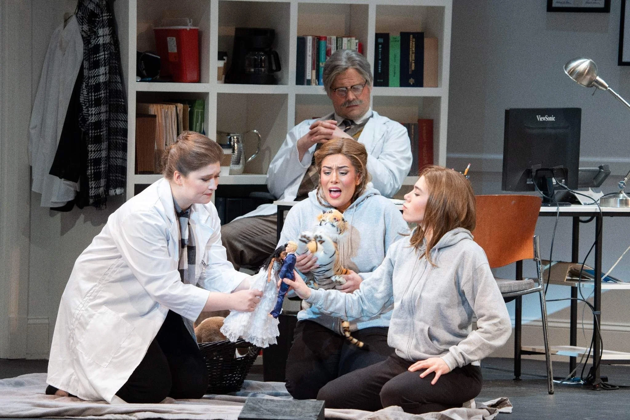 A theatrical scene featuring four actors engaging with puppets in a doctor’s office or therapy setting, with a bookshelf and office equipment in the background.