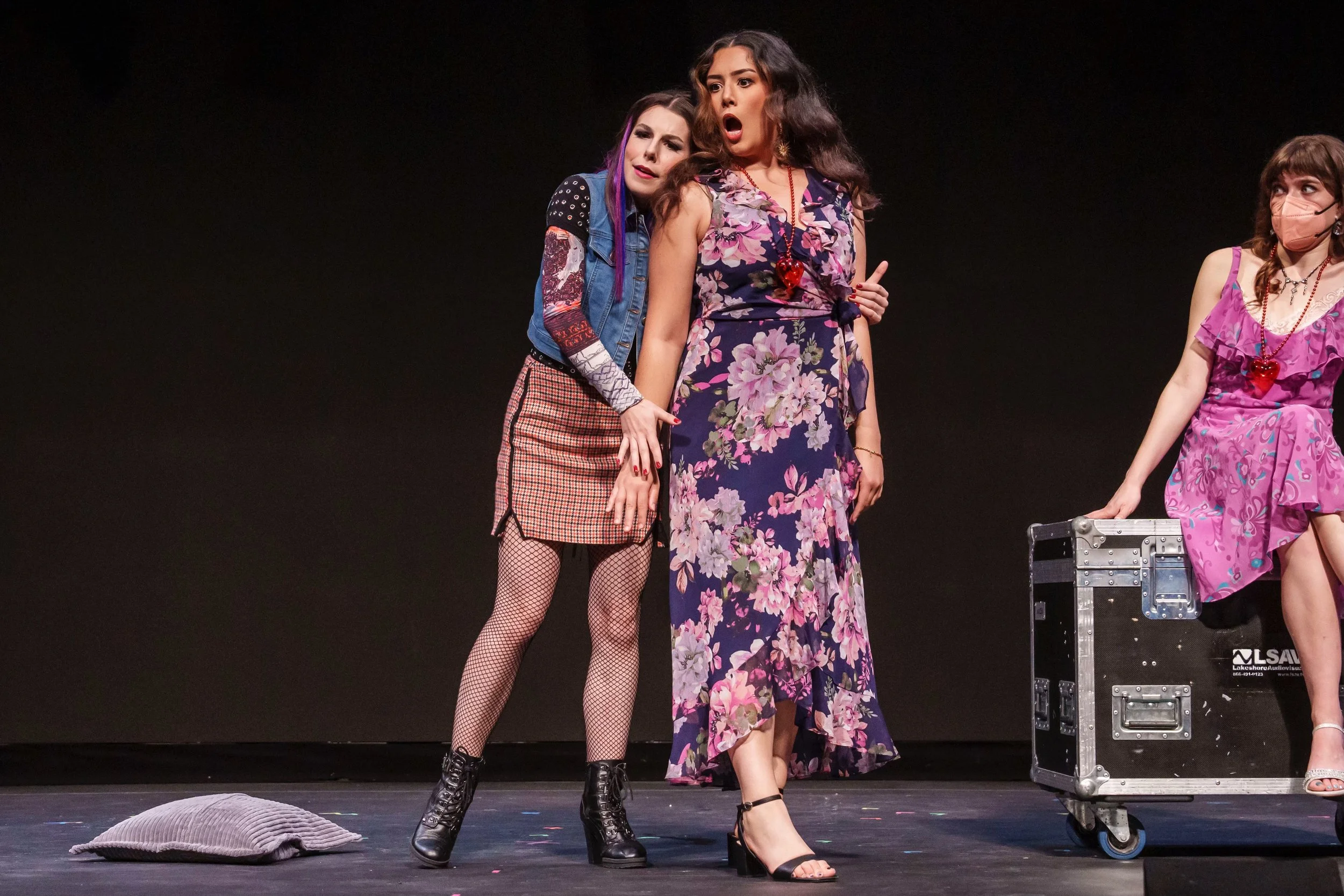Three women on stage, two of whom are dressed in floral dresses and one in a casual outfit with a patchwork skirt and fishnet stockings, appear to be acting or performing. There is a black backdrop, a pillow on the floor, and a case on wheels.