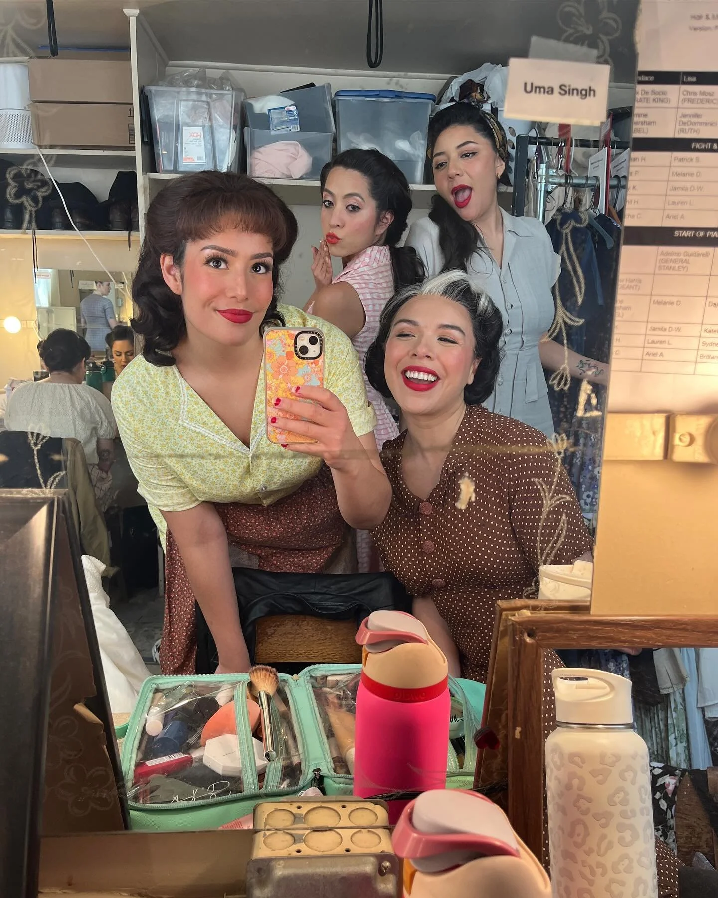 A group of five women in vintage clothing taking a selfie in front of a mirror, with makeup and hair styling items visible on the vanity, and a dressing room background.