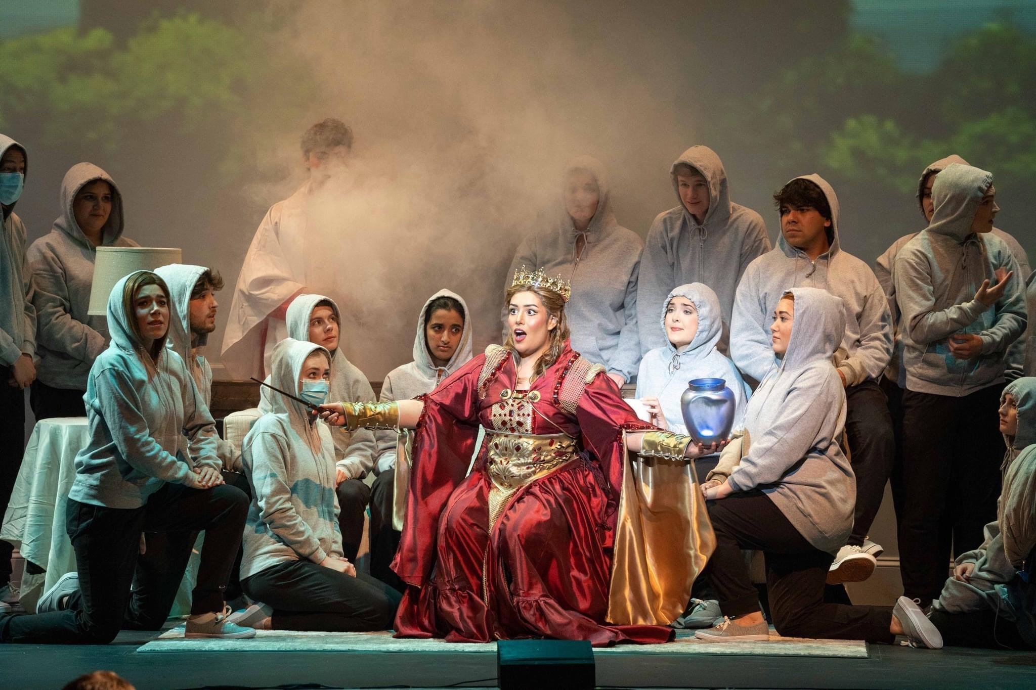 A theatrical performance featuring a woman dressed as a queen in a red gown and crown, seated on stage surrounded by cast members in casual hoodies and masks. The scene includes stage fog and a backdrop of trees.