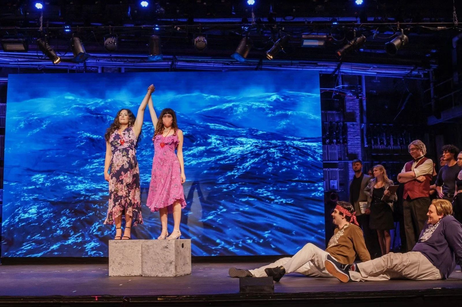 The stage is set with a blue ocean backdrop. Two women are standing on a platform at the center, holding hands raised in victory or farewell. They are dressed in floral dress, one in purple and the other in pink. Several people are seated and standin