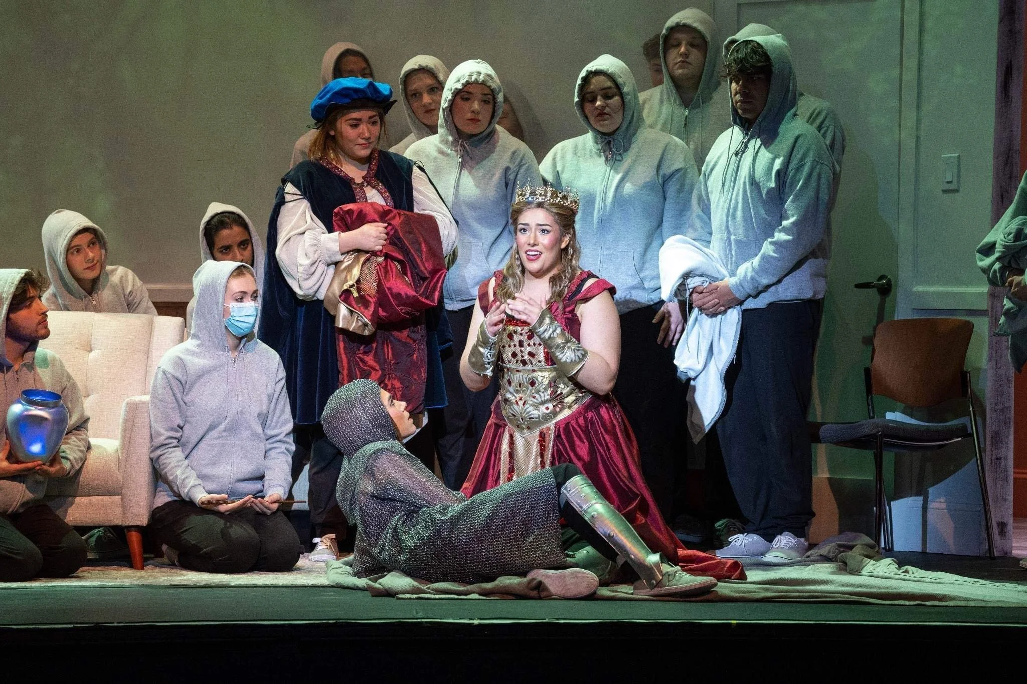 Actors performing on stage, with a woman dressed as a queen sitting on the ground and others gathered around her, some wearing costumes and casual clothing.