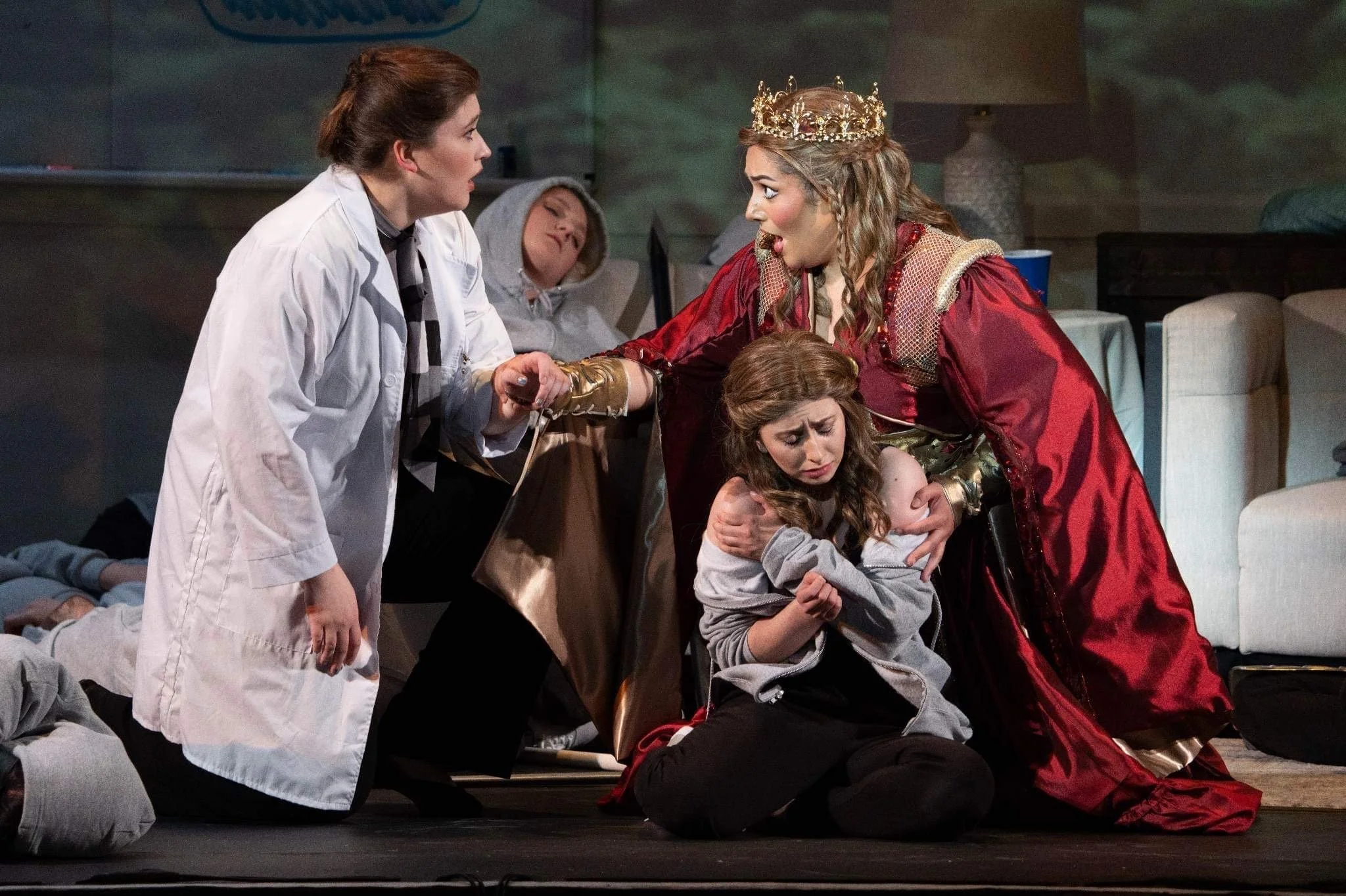 The image shows a dramatic scene on a stage, with several women and children. A woman in a white coat and checkered scarf is kneeling and speaking passionately to a woman dressed as a queen, who is wearing a crown and an elaborate red gown with gold 