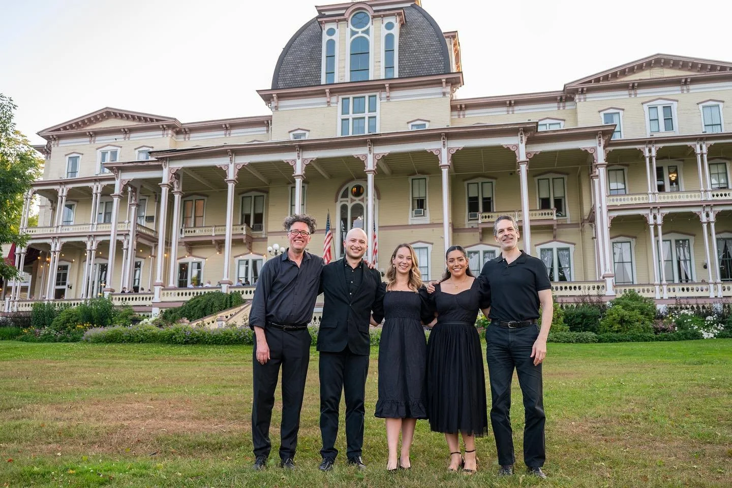Five singers standing on lawn in front of a large, ornate Victorian-style house, smiling at the camera, with greenery and flowers in the garden.