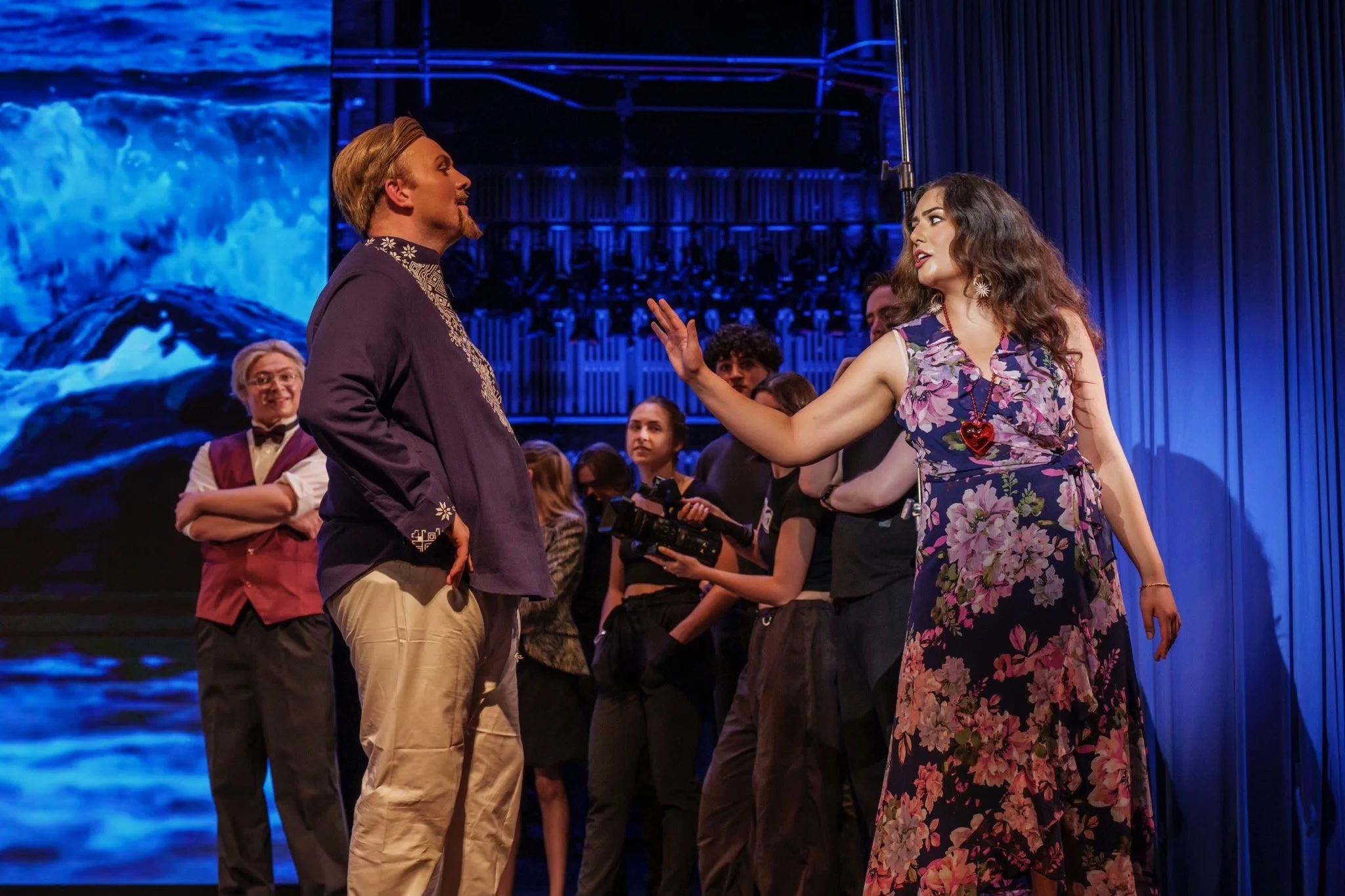 A woman in a floral dress and red necklace gestures toward a man in a purple embroidered shirt on stage, with several people and a blue background behind them.