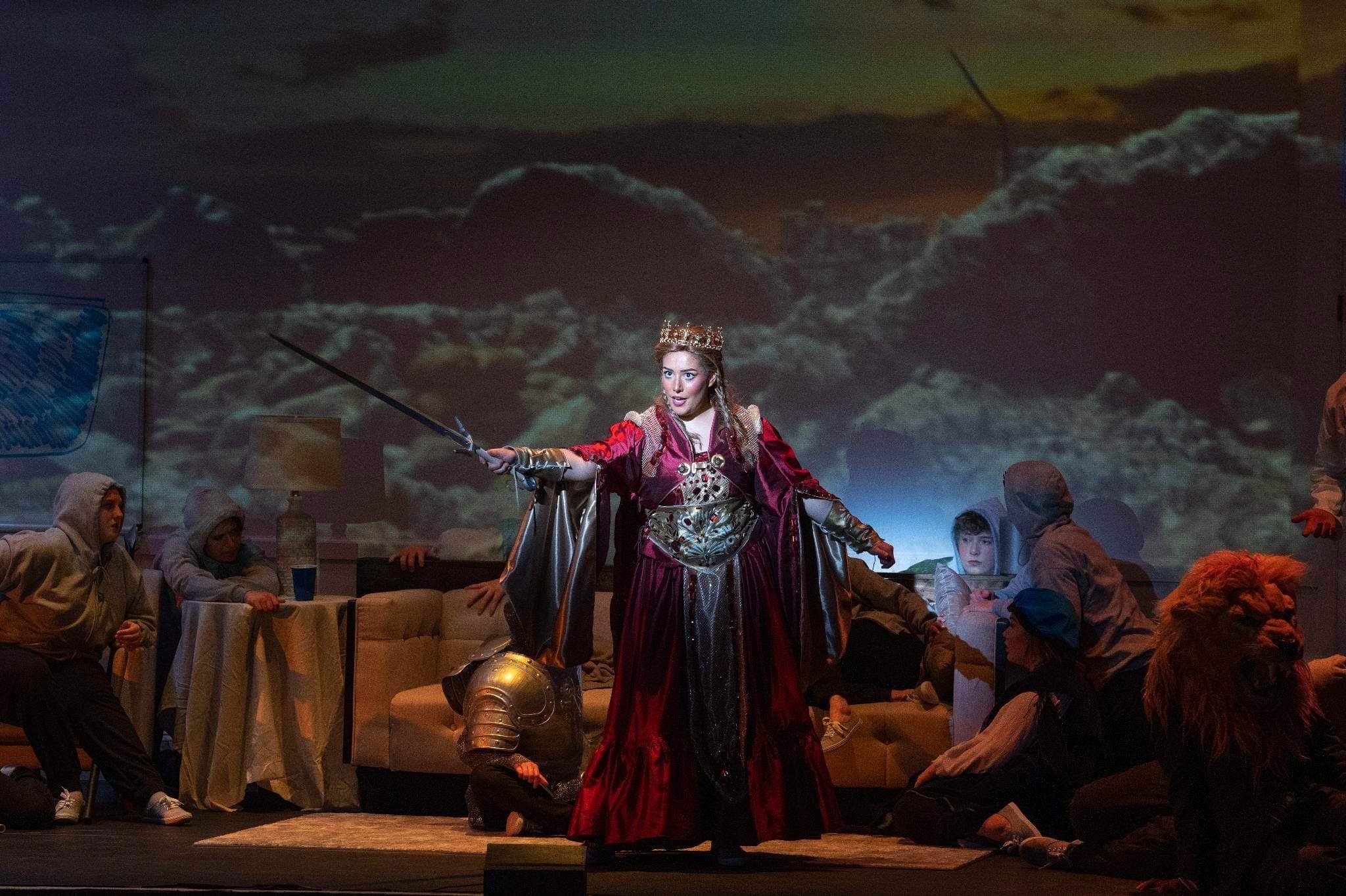 Stage scene from a theatrical performance featuring a woman dressed as a queen in a red and gold costume with a crown, holding a sword. She stands with an intense expression, surrounded by actors wearing hooded costumes and animal masks, with a backd