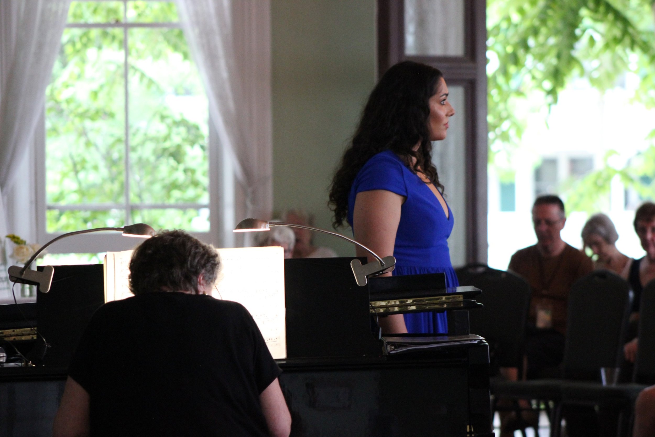 Soprano Uma Singh in a blue dress standing near a piano during a performance at Central City Opera, with an audience in the background.