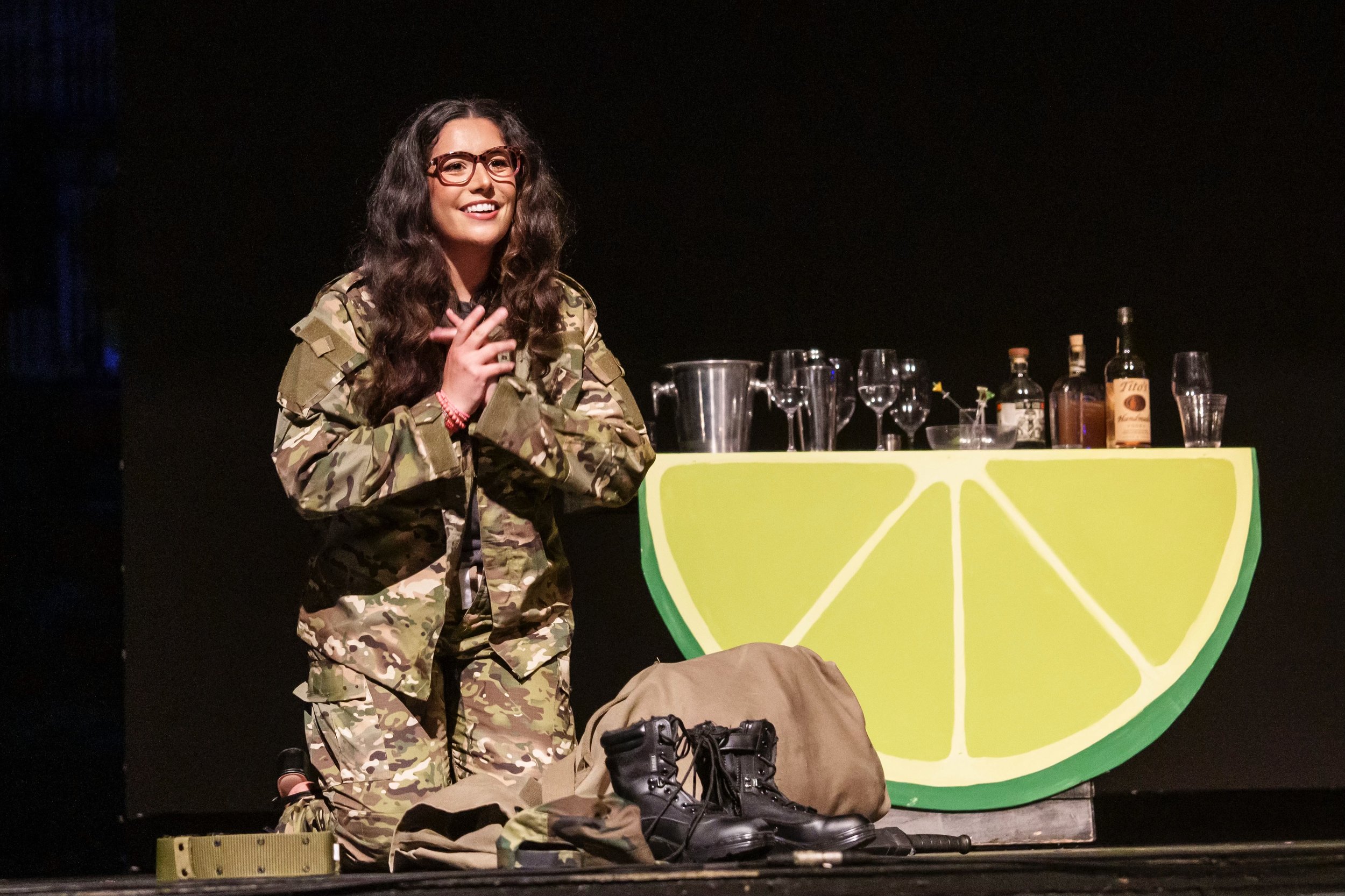 Young woman with glasses in military camouflage uniform standing on stage next to a lime wedge-shaped bar with various bottles and glasses on top. There are boots, a bag, and other items on the floor in front of her.