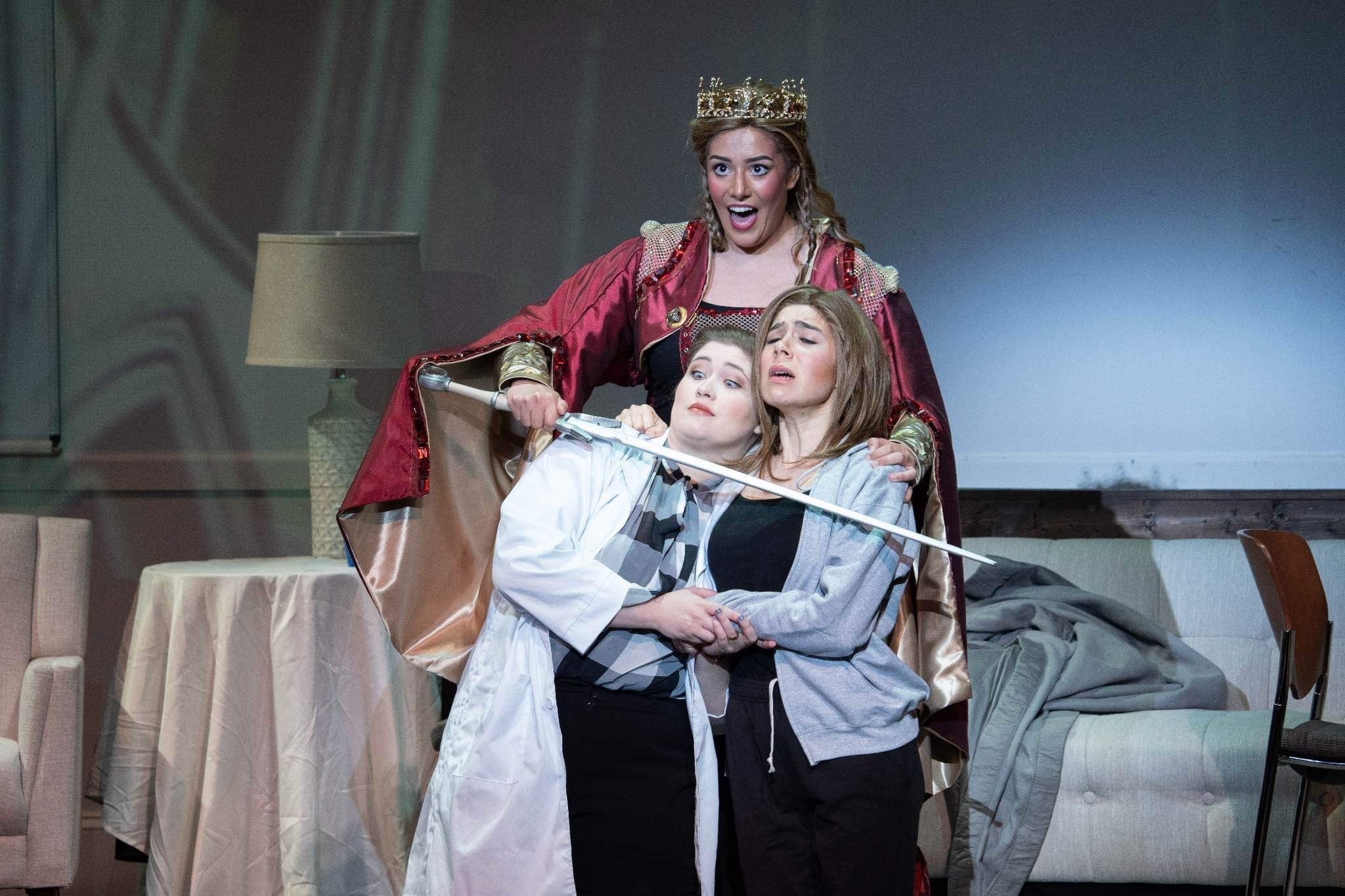 Three young women are on stage, with one dressed as a queen wearing a crown and a royal red robe, her hand on two other women who are dressed casually. They appear to be in a dramatic scene, with the woman dressed as a queen looking surprised or exci