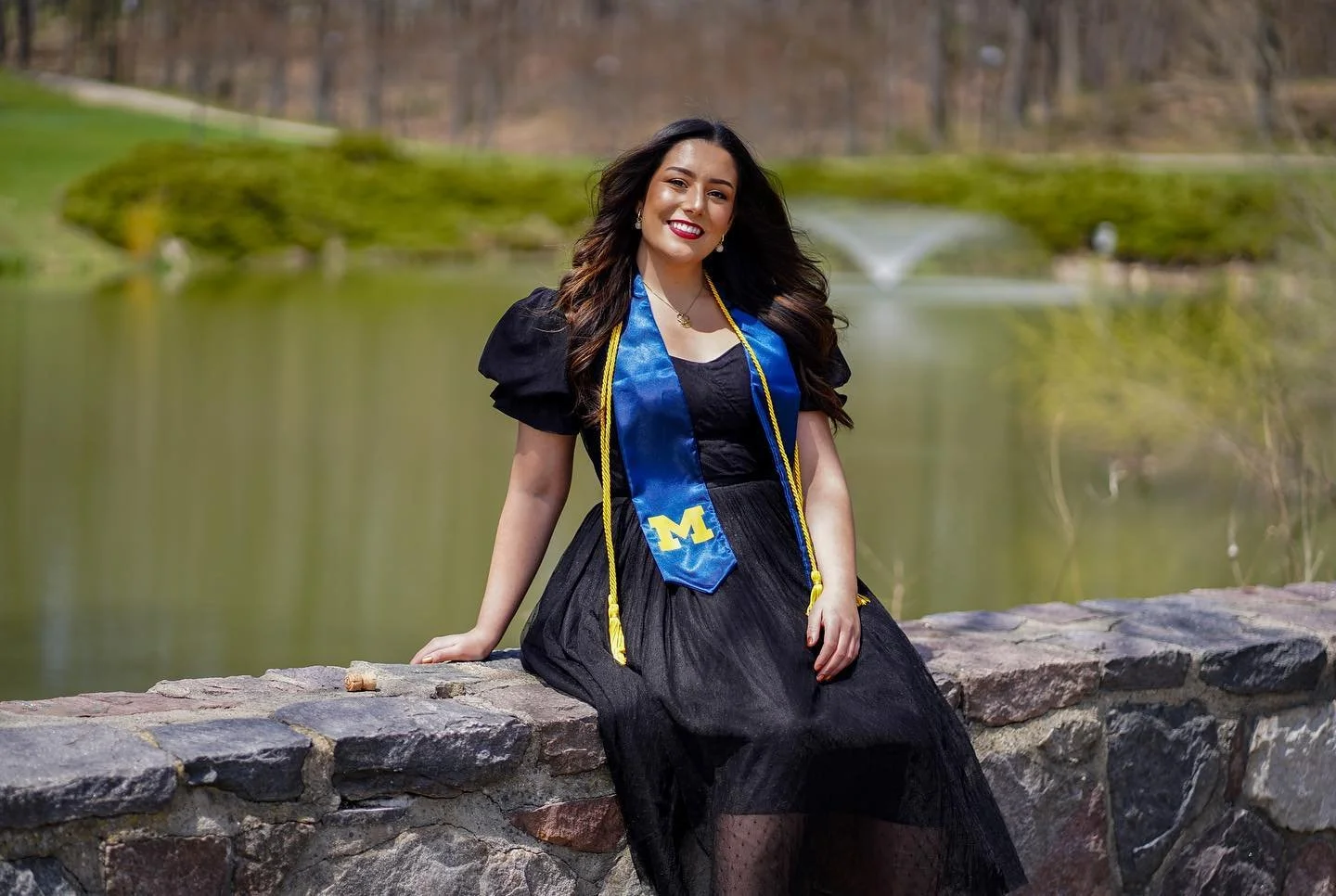 Uma Singh graduating from the University of Michigan, sitting on a stone wall by a lake with trees and a fountain in the background.